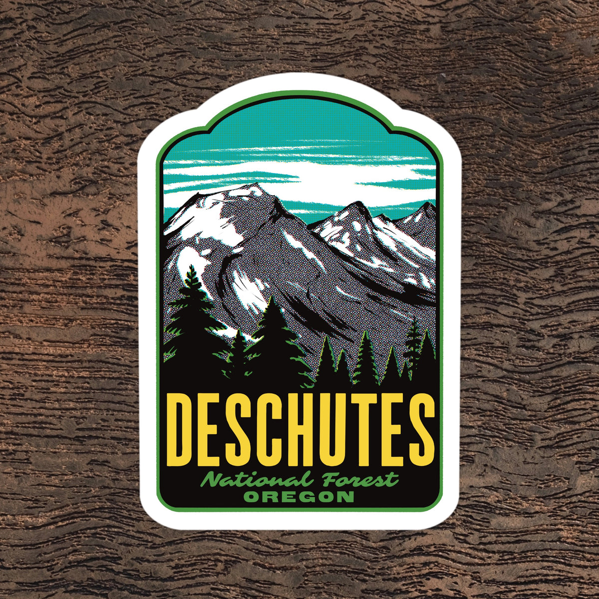 A die-cut sticker featuring a stylized mountain landscape with pine trees and the text 'DESCHUTES National Forest OREGON,' shown on a dark wood background.