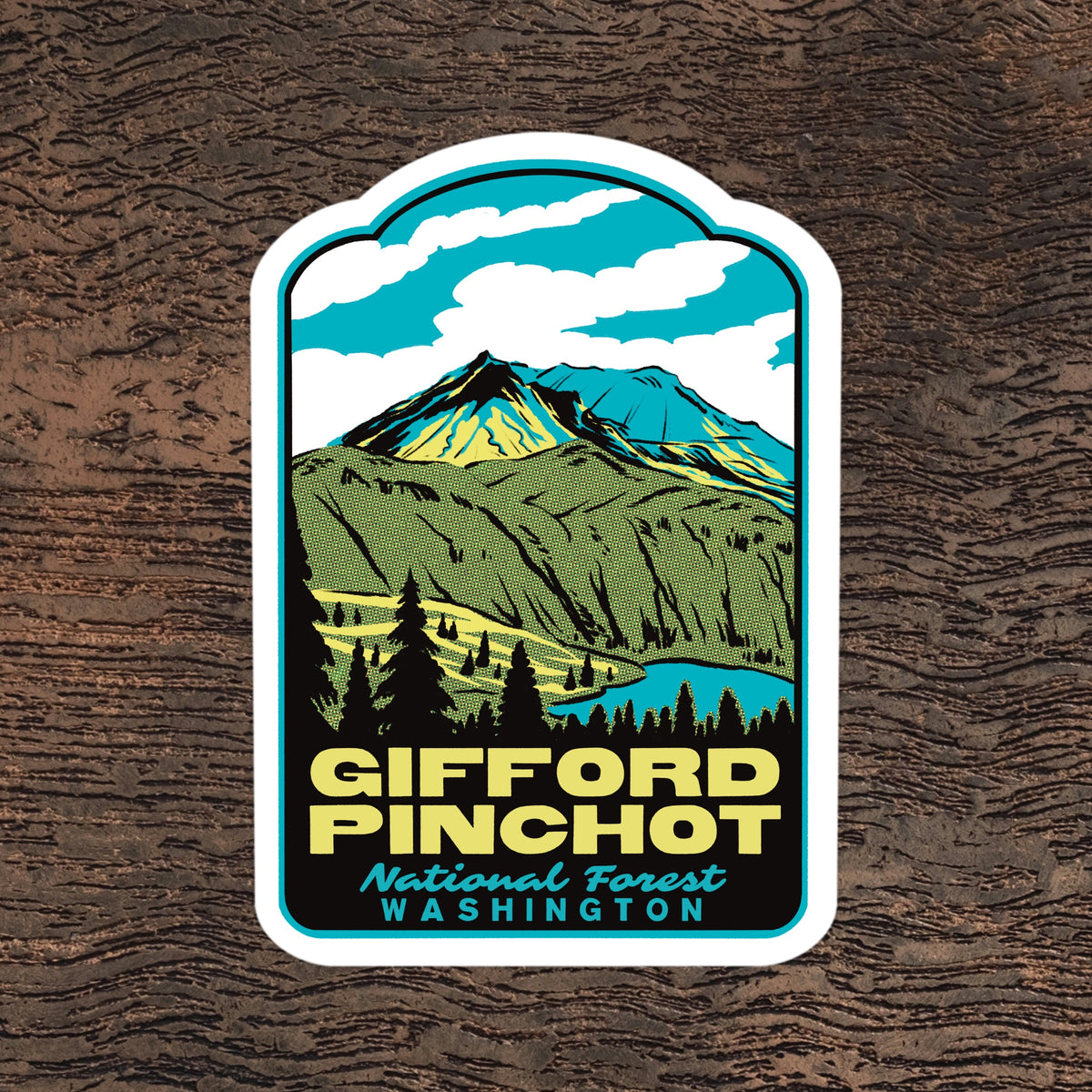 A die-cut sticker featuring a stylized mountain landscape with the text 'Gifford Pinchot National Forest Washington,' shown on a dark wood background.