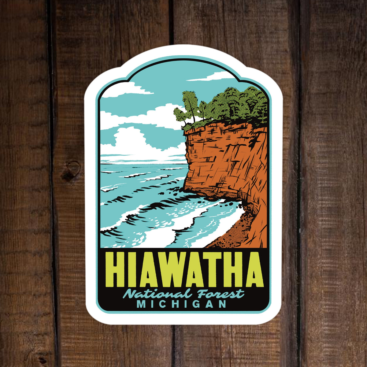 A die-cut sticker featuring a retro-style illustration of the Hiawatha National Forest in Michigan, with blue water, white clouds, green trees, and an orange-brown cliff, displayed on a dark wood background.