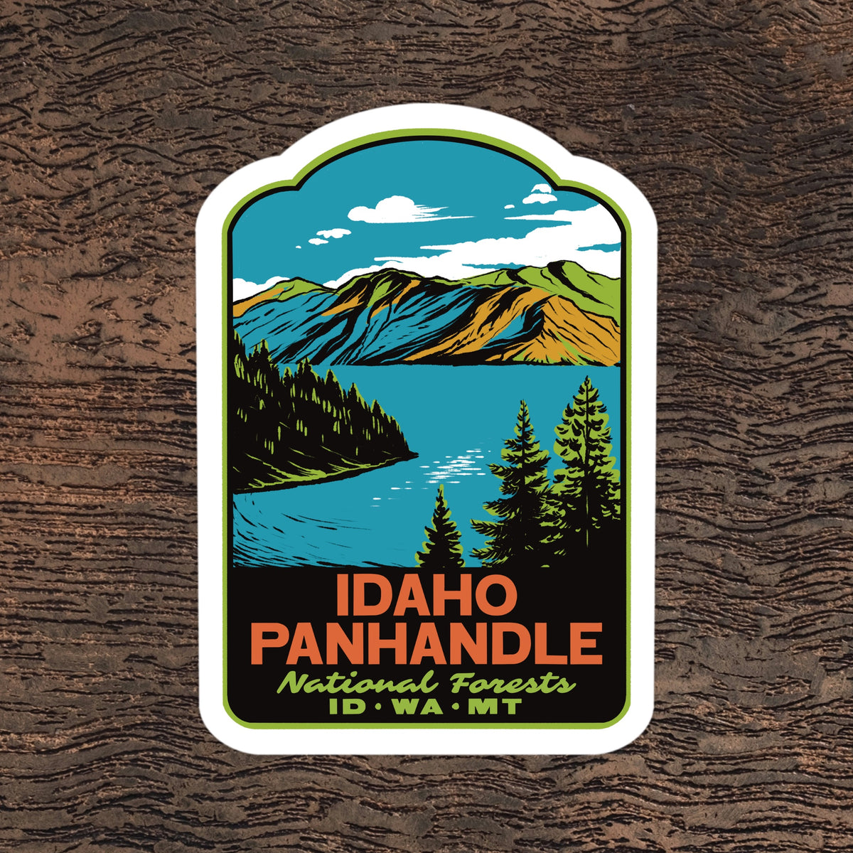 A die-cut sticker featuring a stylized illustration of Idaho Panhandle National Forests, including mountains, a lake, pine trees, and the text 'IDAHO PANHANDLE National Forests ID·WA·MT', shown on a dark wood background.