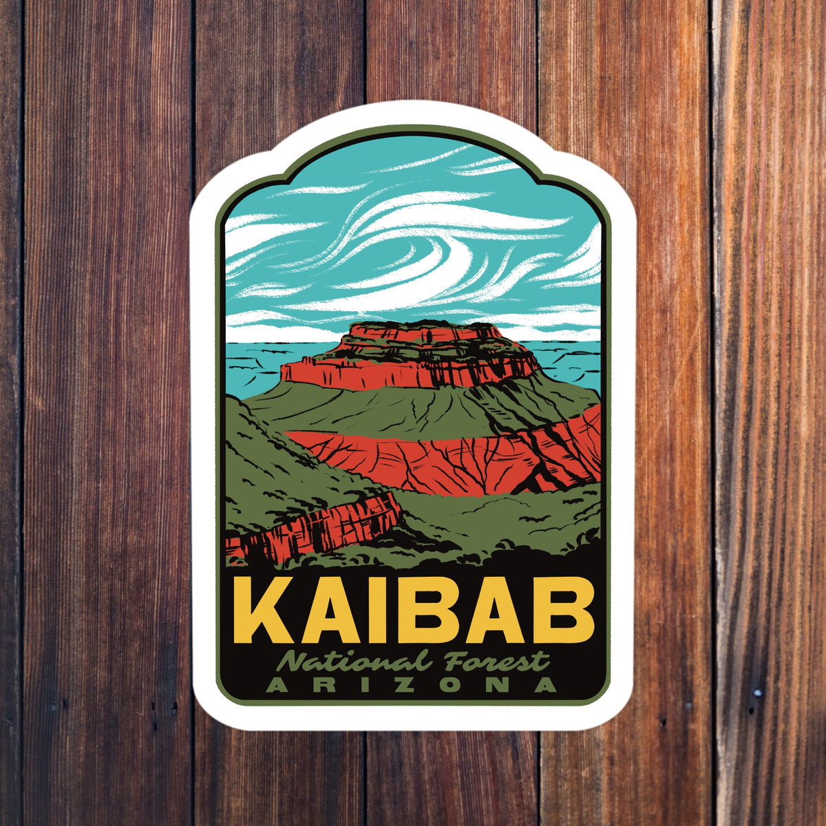 A kiss-cut sticker featuring a retro-style illustration of Kaibab National Forest in Arizona, with layered red rock mesas, green hills, a turquoise sky, and bold yellow text reading 'KAIBAB' and smaller text 'National Forest ARIZONA', shown against a dark wood plank background.