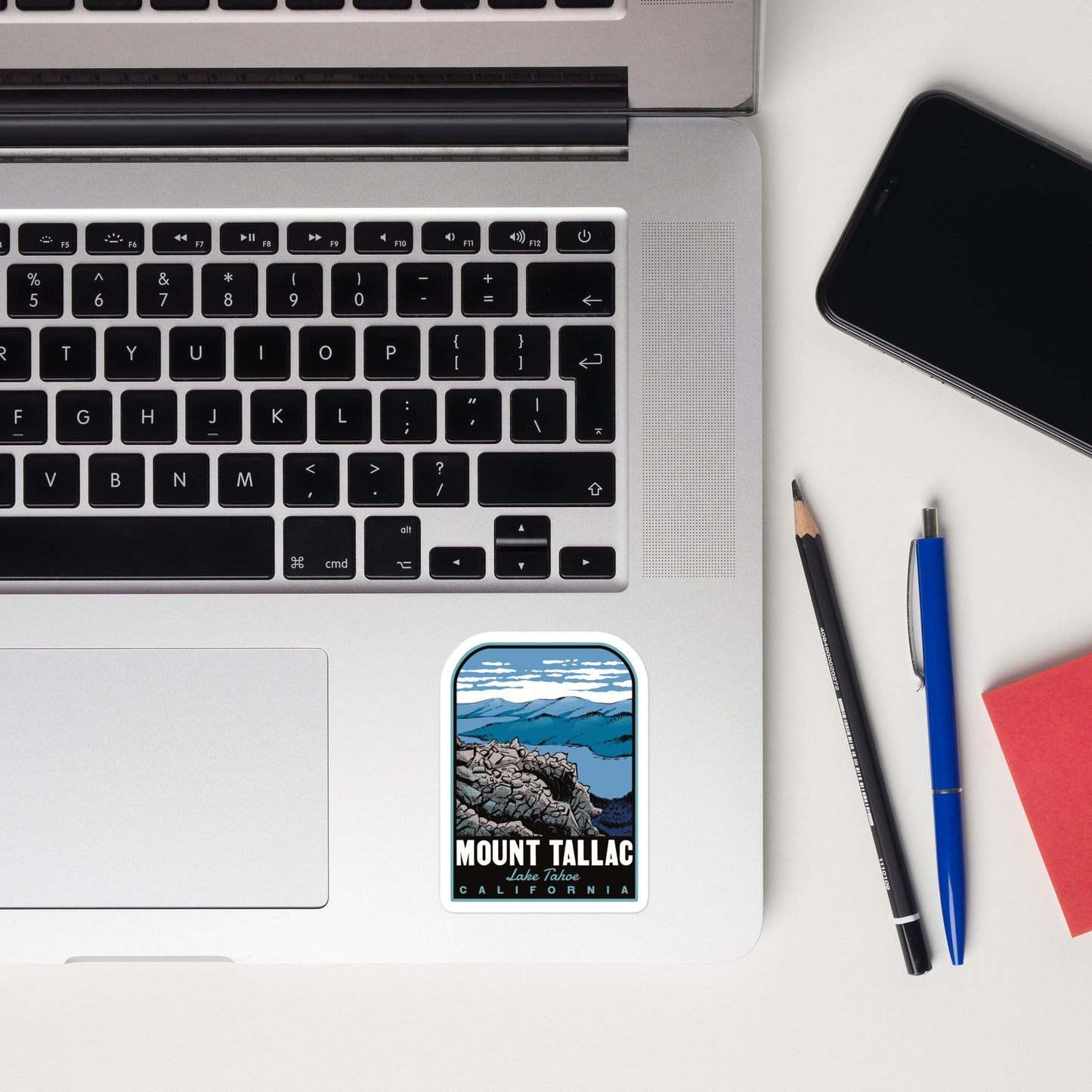 Laptop with a sticker of Mount Tallac, California, next to a phone, pencil, and pen on a white surface.