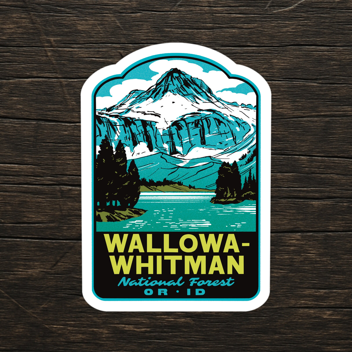 A die-cut sticker featuring a retro-style illustration of Wallowa-Whitman National Forest, with a turquoise mountain, lake, and trees, and bold yellow text reading 'WALLOWA-WHITMAN National Forest OR ID' on a dark wood background.