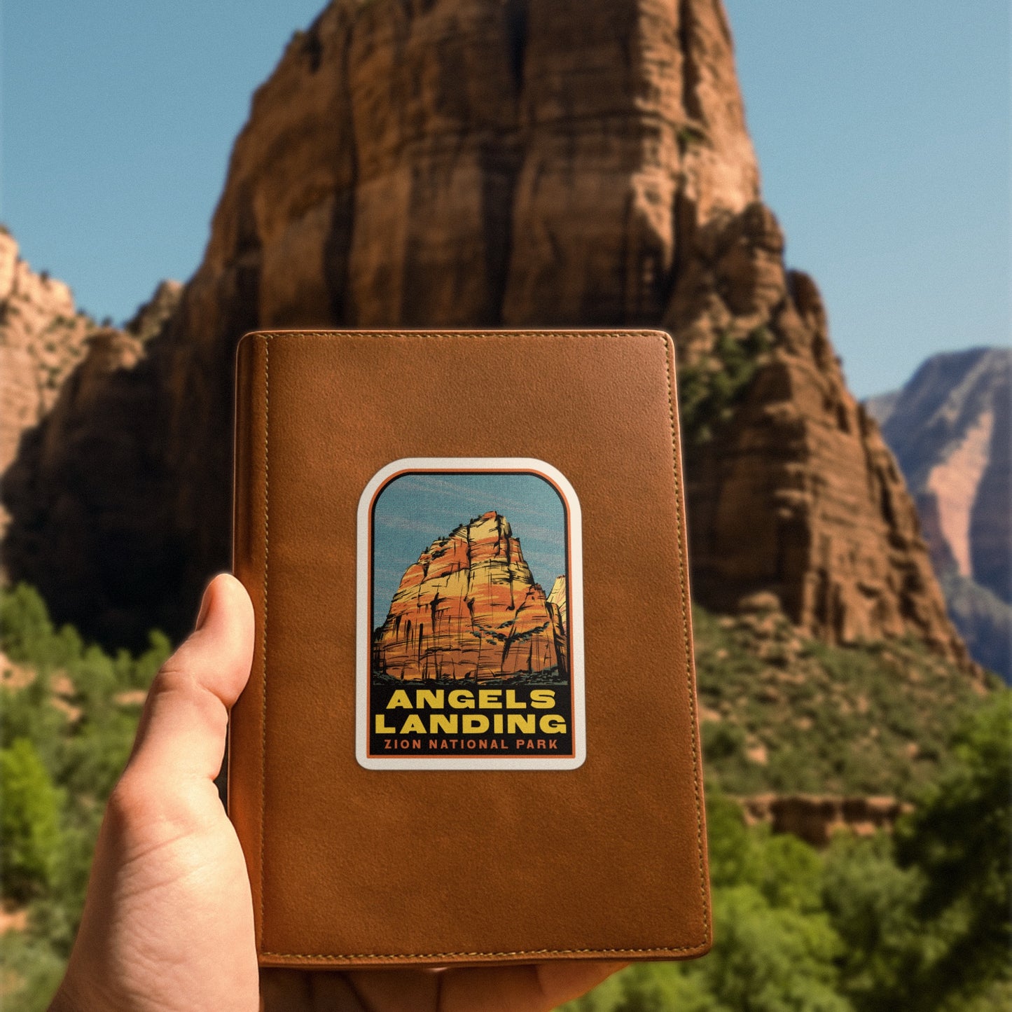Brown leather wallet with Angels Landing Zion National Park patch held in front of a mountainous landscape.