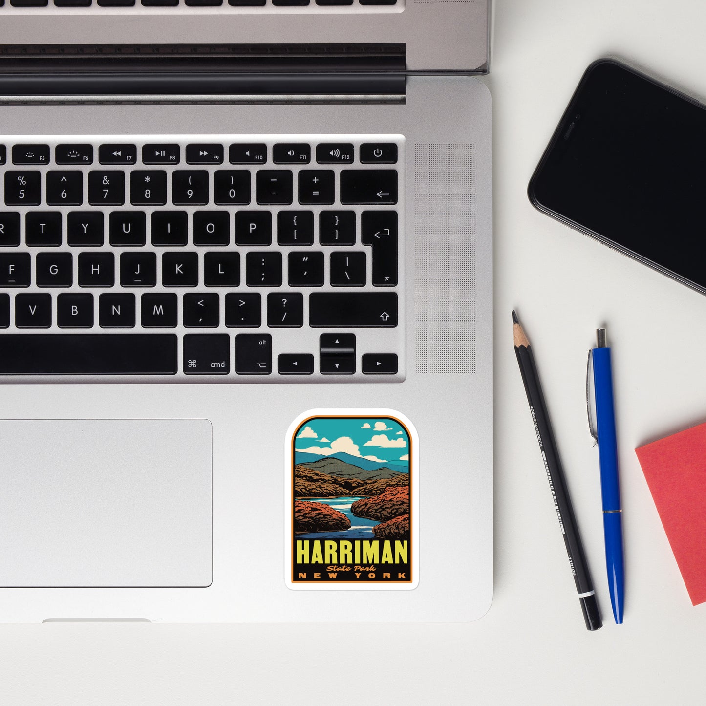 Laptop with a scenic sticker labeled 'Harriman New York', a phone, pencil, and pen on a white surface.