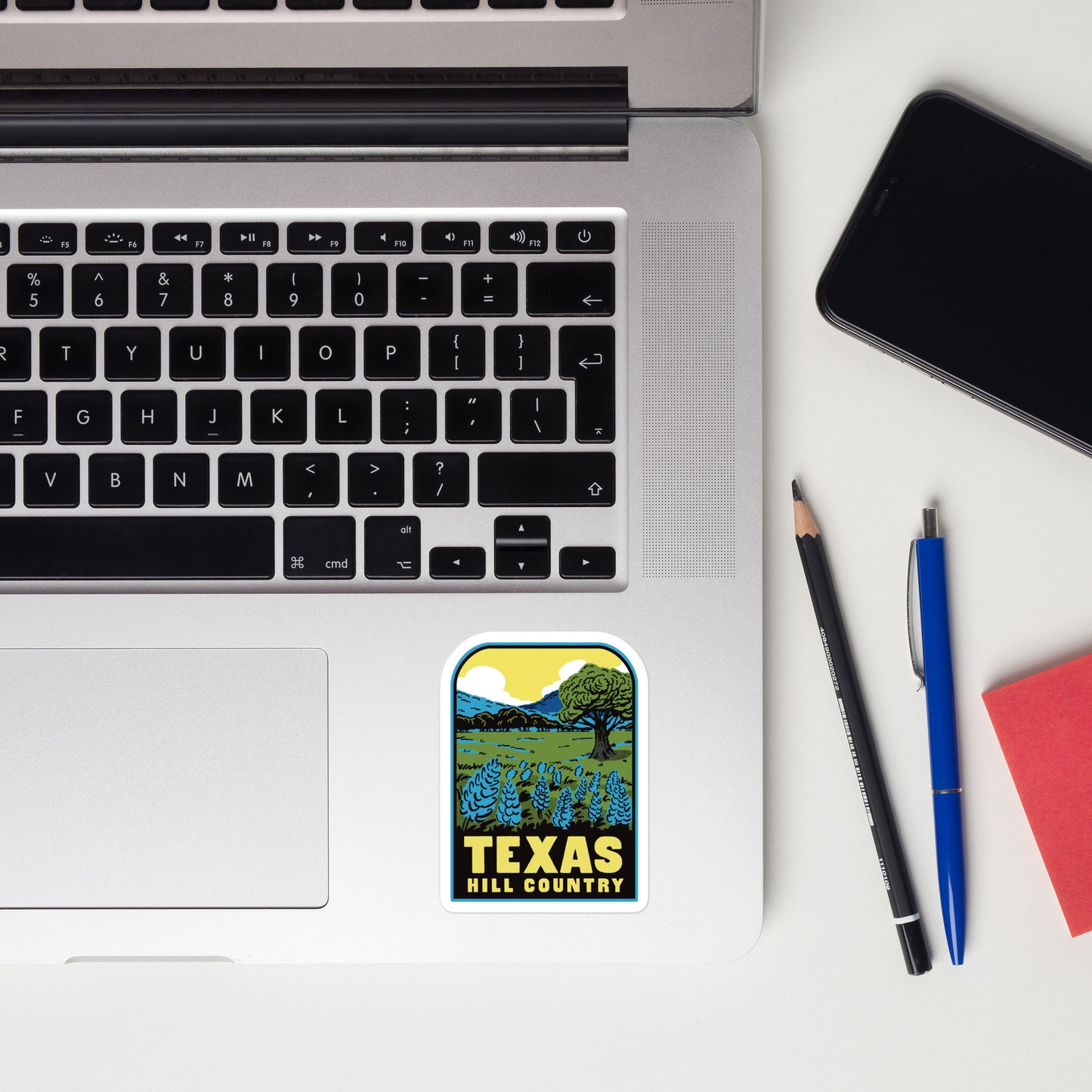 Laptop with a 'Texas Hill Country' sticker, phone, pencil, and pen on a white surface