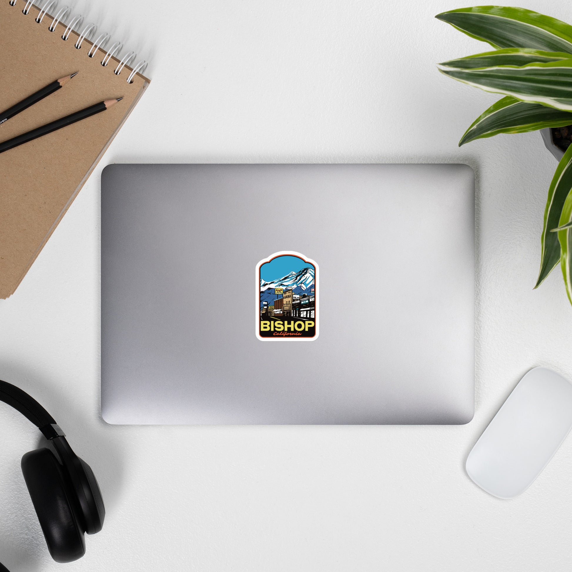 Laptop with a sticker of a Bishop, California on a desk with a notebook, pens, and headphones.