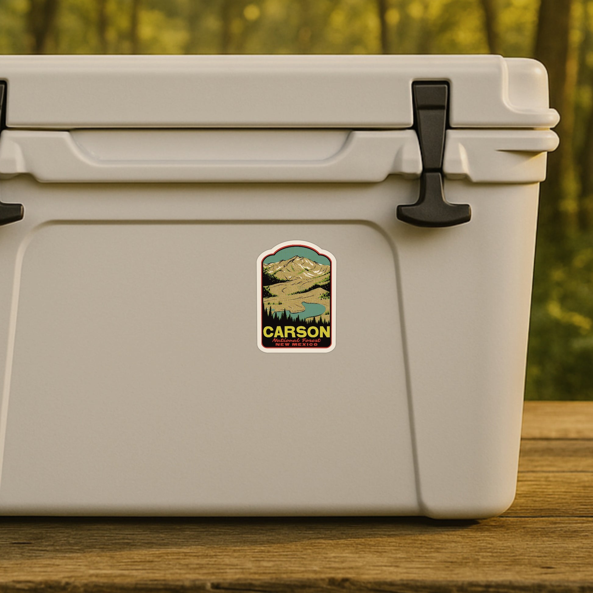 Beige cooler box with a Carson National Forest logo on a wooden surface and blurred natural background
