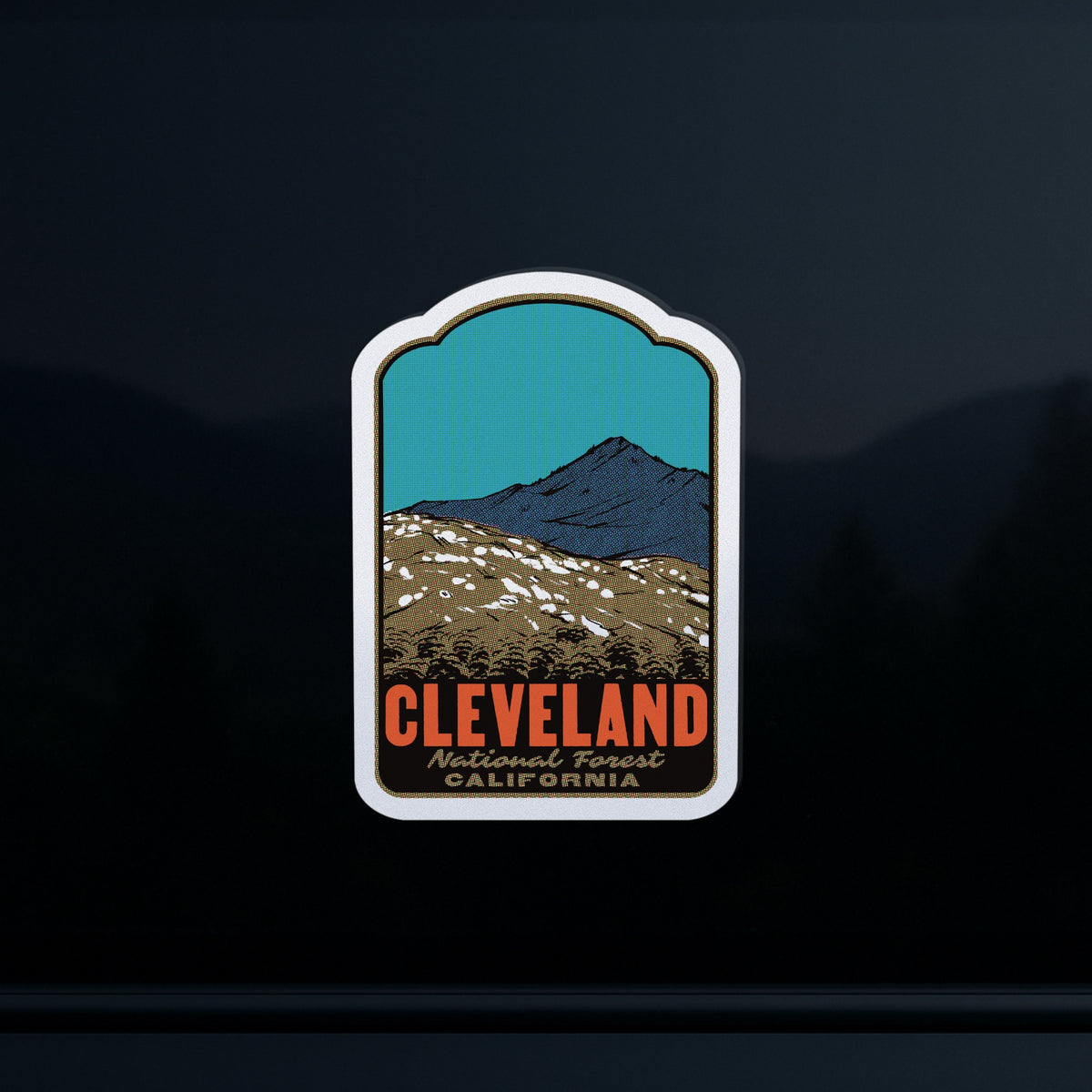 A die-cut sticker featuring an illustrated mountain landscape with the text 'CLEVELAND National Forest CALIFORNIA' in orange and black, shown on a dark reflective surface.