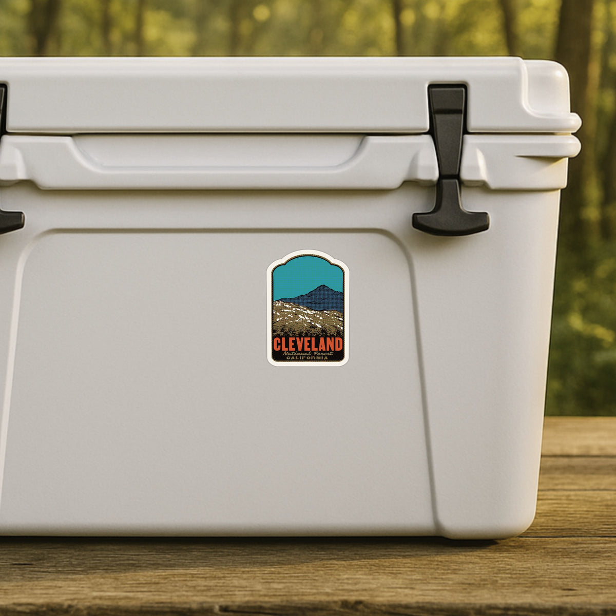A white cooler sitting on a wooden surface outdoors, with a small Cleveland-themed sticker on the front and a blurred green forest background.