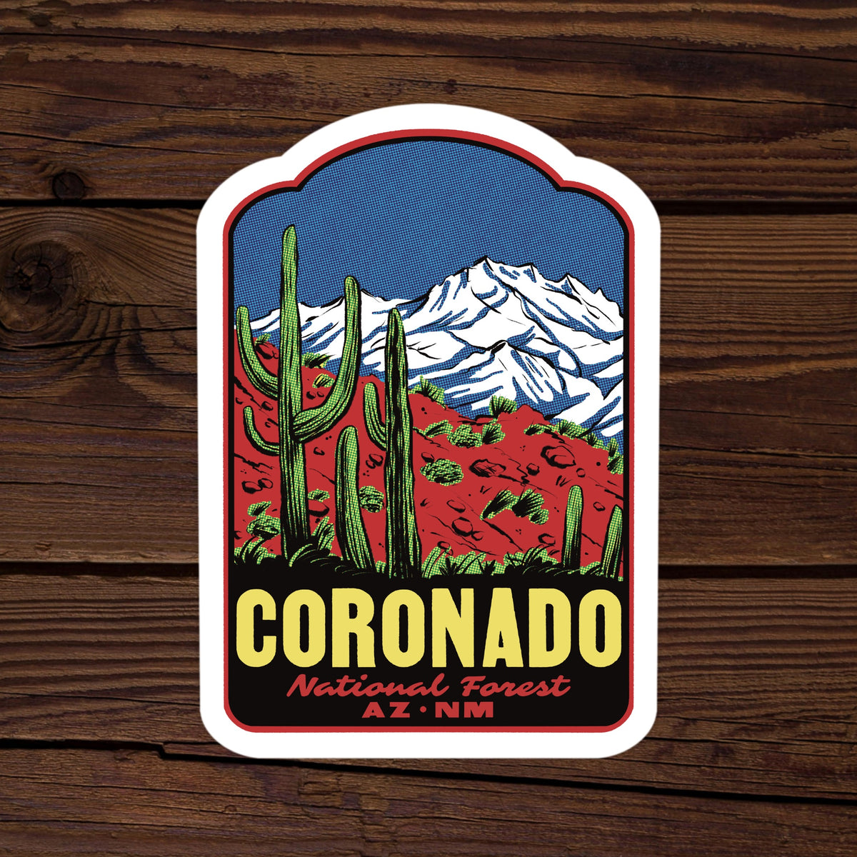 A die-cut sticker featuring a stylized illustration of Coronado National Forest with saguaro cacti, red desert terrain, and snow-capped mountains under a blue sky. The sticker reads 'CORONADO National Forest AZ NM' and is shown on a dark wood plank background.