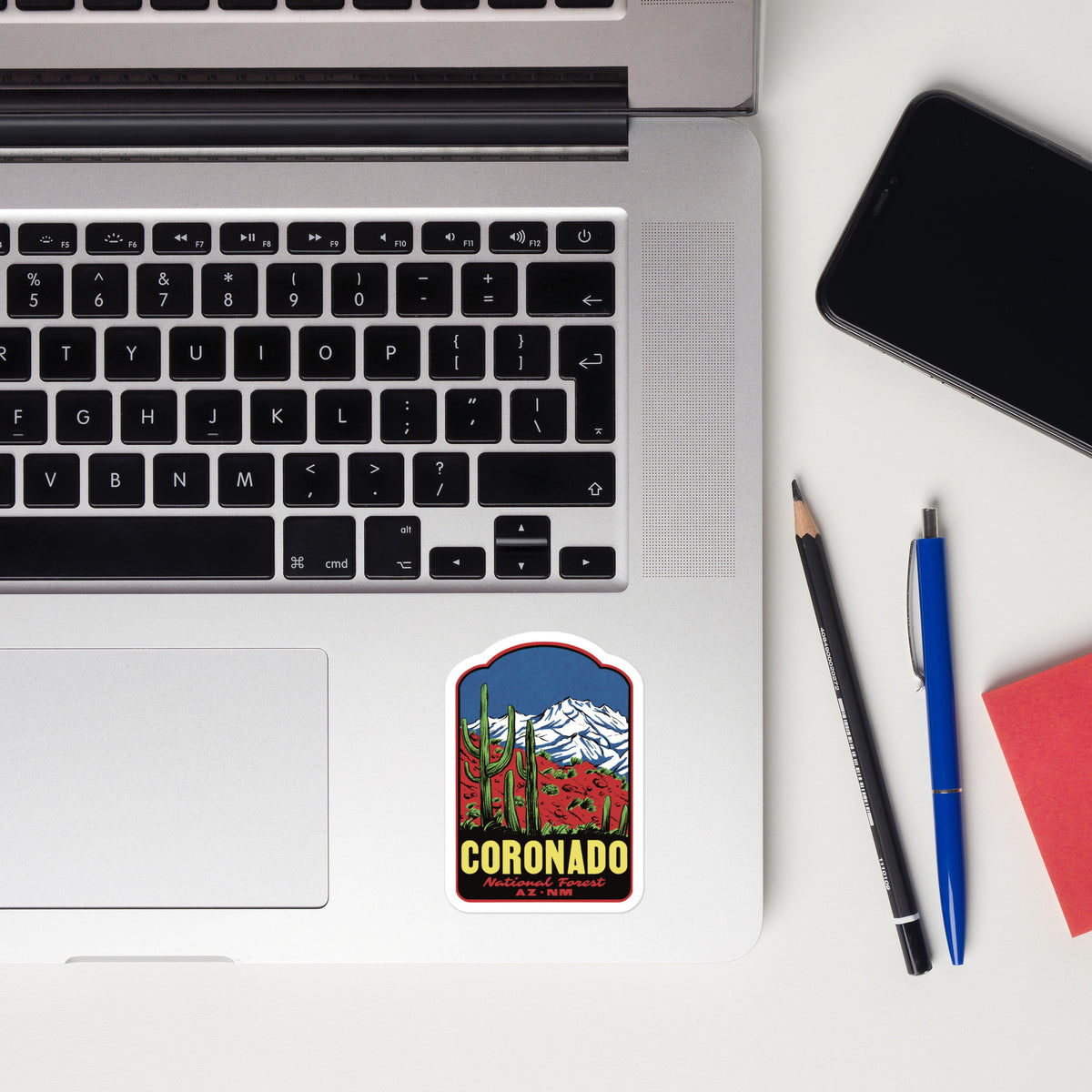 A rectangular sticker with rounded corners is placed on the lower part of a silver laptop keyboard. The sticker features a colorful illustration of saguaro cacti, red flowers, and a snow-capped mountain under a blue sky, with the word 'CORONADO' in yellow at the bottom.