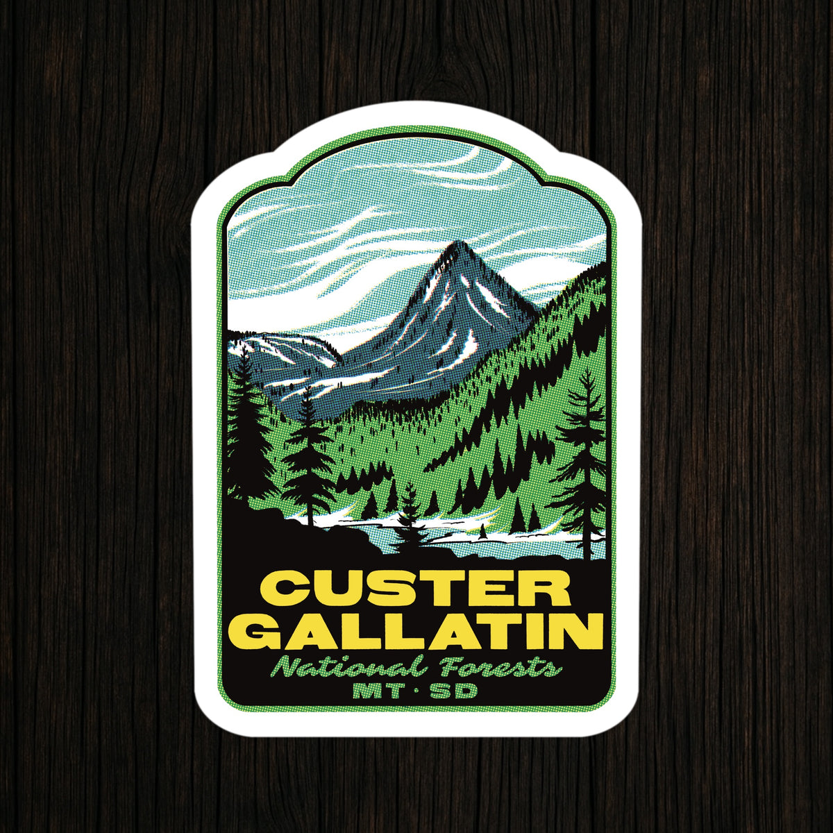 A die-cut sticker featuring a stylized mountain landscape with pine trees and the text 'CUSTER GALLATIN National Forests MT. SD', shown on a dark wood background.