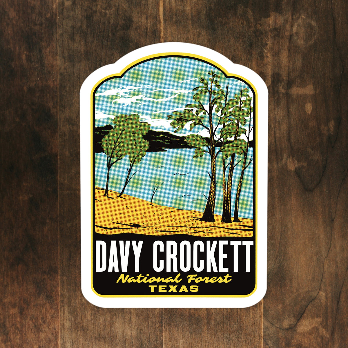 A die-cut sticker featuring a vintage-style illustration of Davy Crockett National Forest in Texas, with trees, blue water, and a sandy shore, placed on a dark wood background.