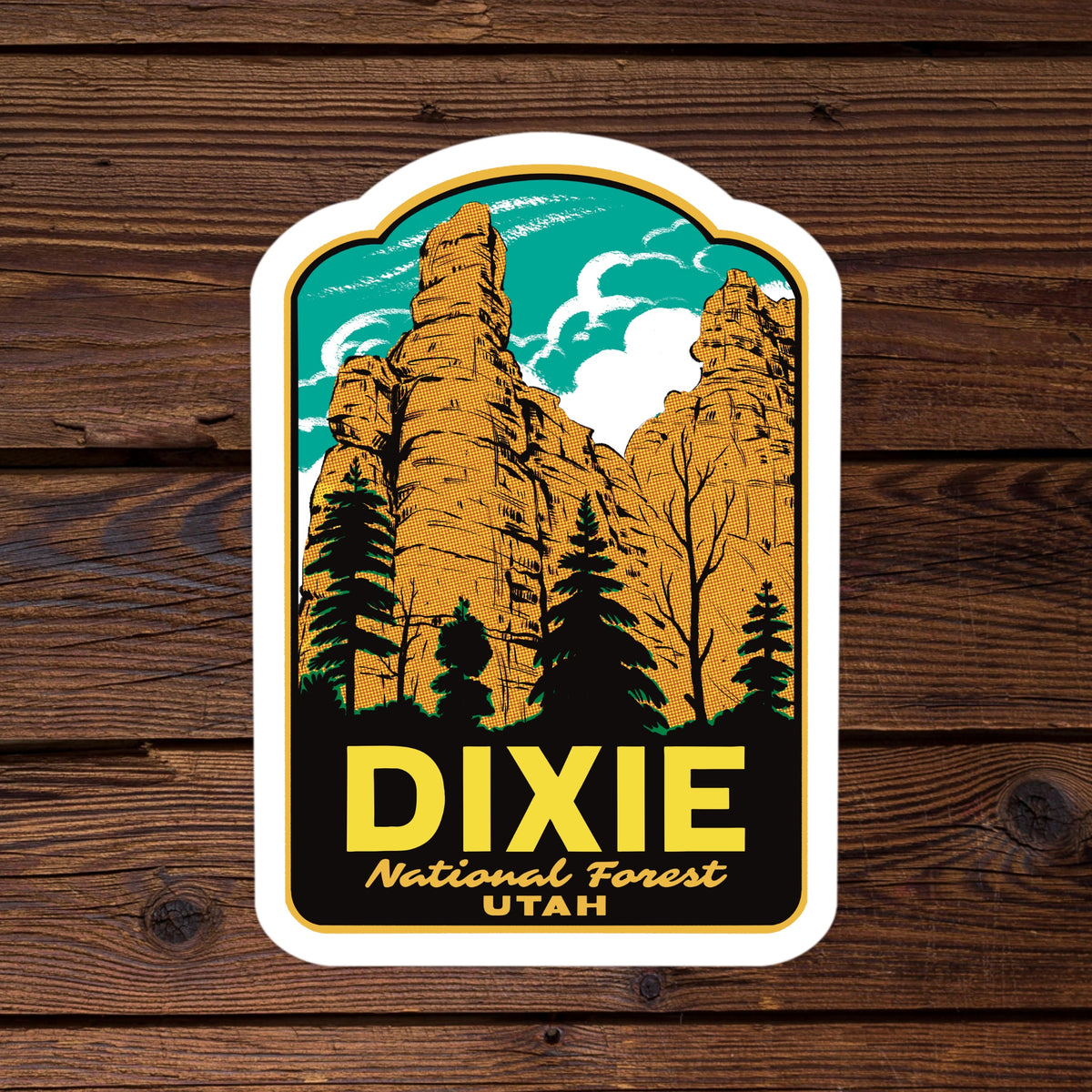 A die-cut sticker featuring a retro-style illustration of Dixie National Forest in Utah, with tan rock formations, dark green trees, and a teal sky, displayed on a dark wood plank background.