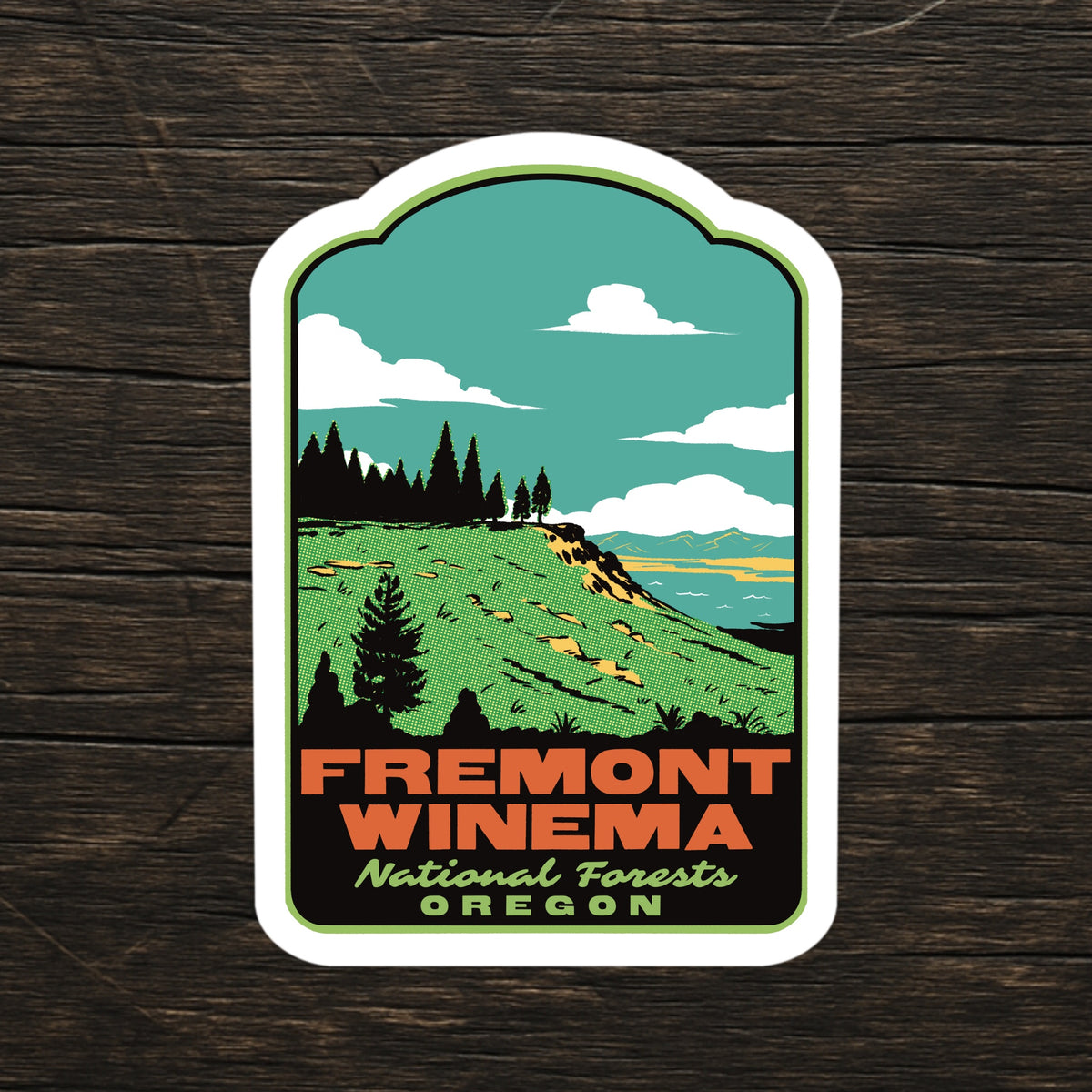 A die-cut sticker featuring a retro-style illustration of Fremont Winema National Forests in Oregon, displayed on a dark wood background.