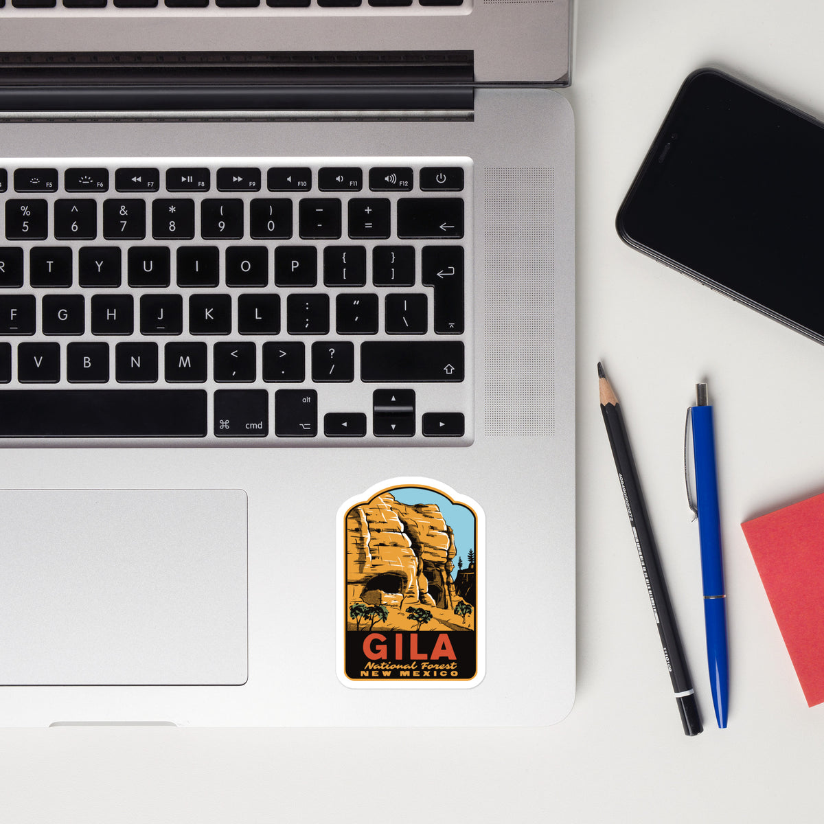 A rectangular sticker with rounded corners featuring a desert rock formation and the text 'GILA NEW MEXICO' is placed on the palm rest of a silver laptop. The laptop is shown in a styled flat-lay on a white desk alongside a smartphone, a pencil, a blue pen, and a red notebook.