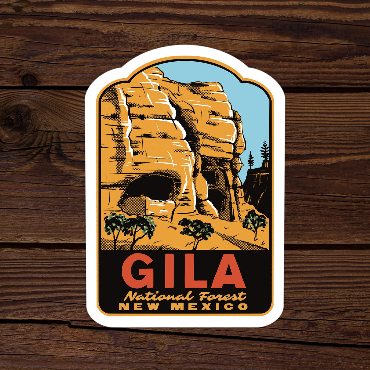 A die-cut sticker featuring a retro-style illustration of Gila National Forest in New Mexico, with a white border, displayed on a dark wood background.
