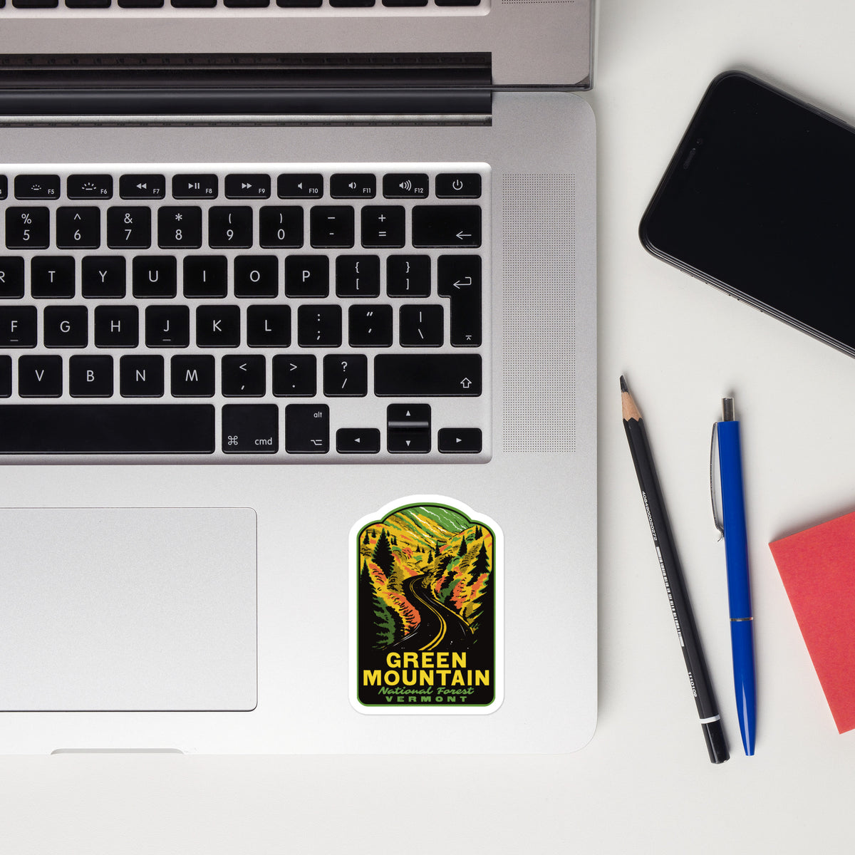 Laptop with a 'Green Mountain National Forest' sticker, phone, pencil, pen, and notebook on a white surface