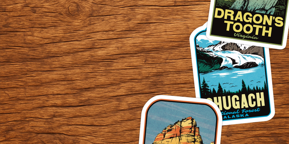 Two vintage-style travel stickers with white borders displayed on a brown wood-grain background. One sticker shows a mountain landscape labeled 'Dragon's Tooth Virginia,' and the other shows a canyon scene labeled 'Chugach National Forest Alaska.
