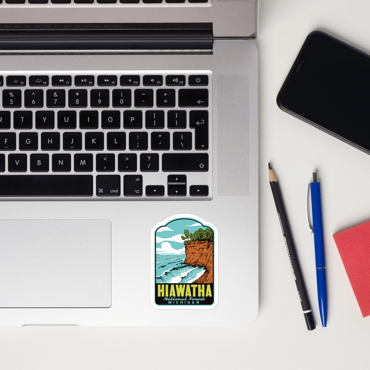 A rectangular sticker with a retro-style illustration of a cliffside beach scene and the text 'HIAWATHA MICHIGAN' is placed on the palm rest of an open silver laptop. The laptop sits on a white desk alongside a black smartphone, a pencil, a blue pen, and a red notebook.