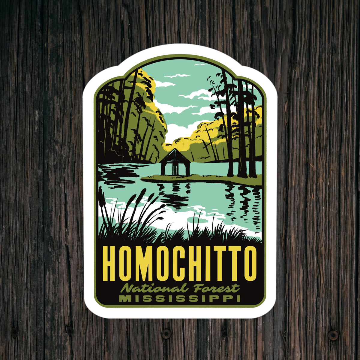 A kiss-cut sticker featuring a retro-style illustration of Homochitto National Forest in Mississippi, with a lake, trees, and a small cabin, displayed on a dark wood background.