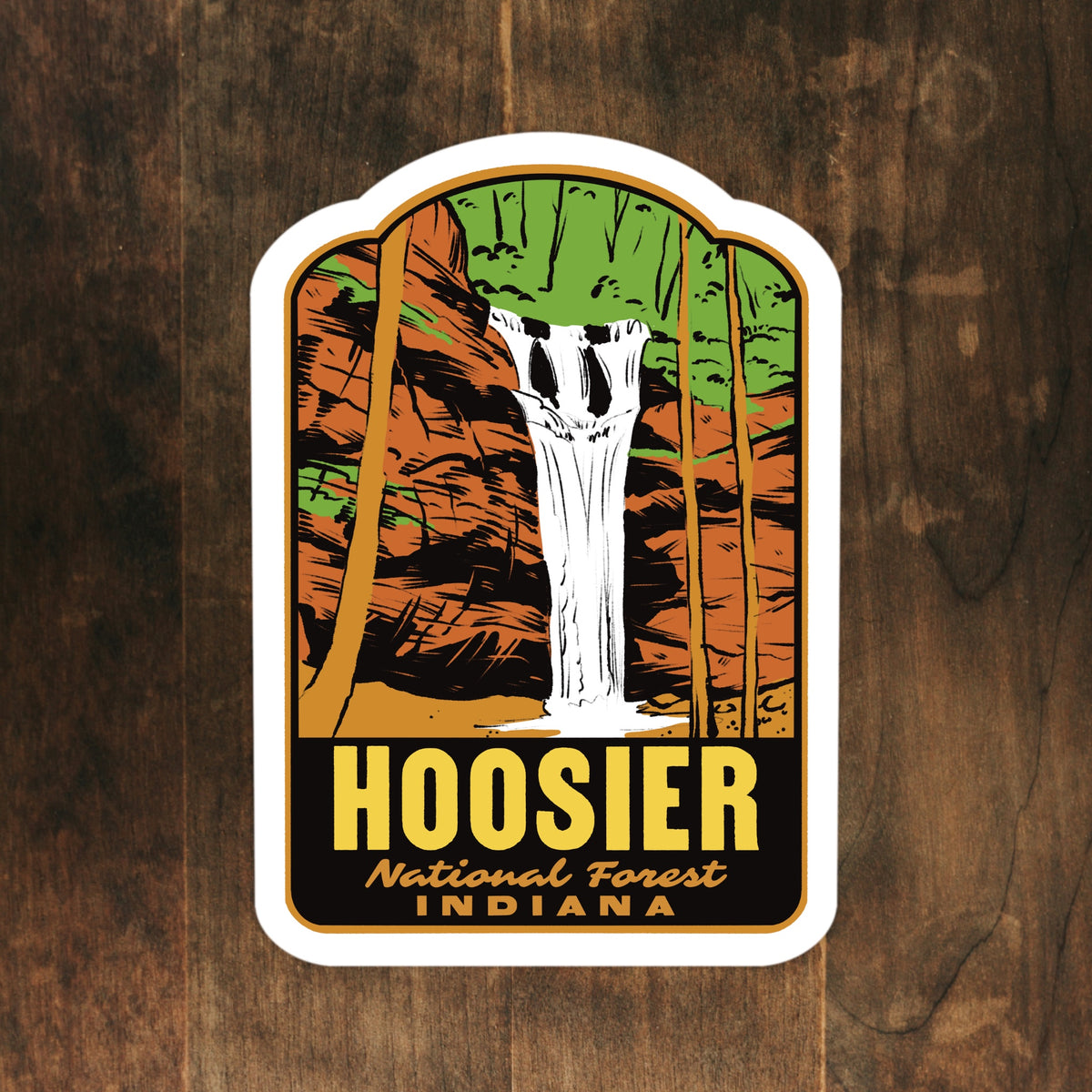 A die-cut sticker featuring an illustration of a waterfall in a forest with the text 'HOOSIER National Forest INDIANA', displayed on a dark wood background.