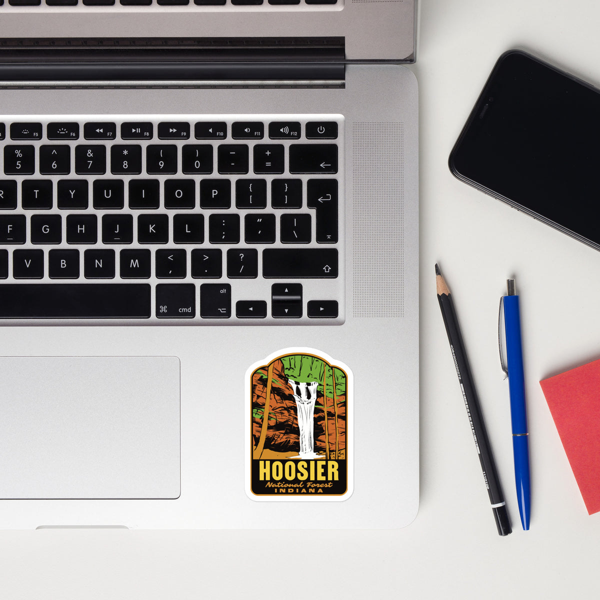 A rectangular sticker with rounded corners placed on the lower left of a silver laptop. The sticker features a stylized illustration of a white skeleton standing in front of a tree with green, orange, and brown leaves, above the word 'HOOSIER' in yellow.