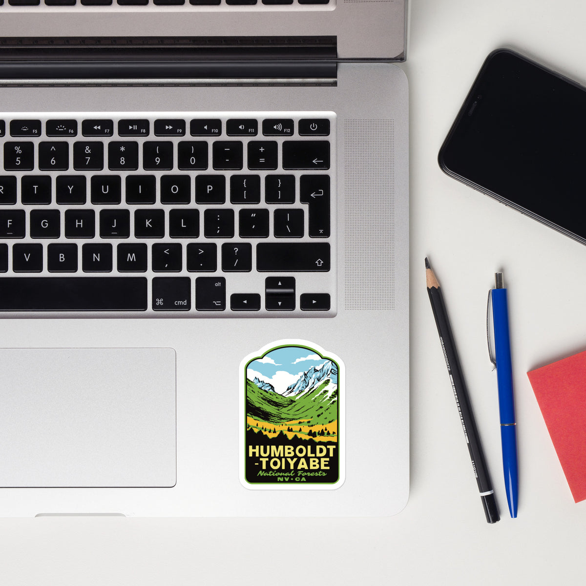A rectangular sticker featuring a mountain landscape and the text 'HUMBOLDT TOIYABE' is placed on the lower left corner of a silver laptop. The laptop is shown in a flat-lay desk setting alongside a smartphone, a pencil, a blue pen, and a red notebook on a white surface.