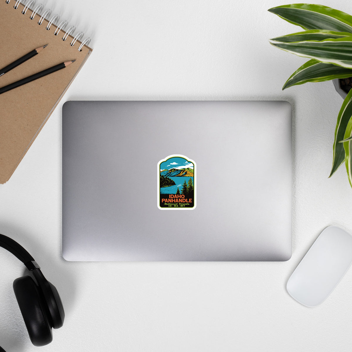 A rectangular sticker with rounded corners is placed on the center of a closed silver laptop. The sticker features a retro-style scenic illustration of a mountain lake with blue sky, green trees, and yellow-orange text reading 'IDAHO PANHANDLE'.