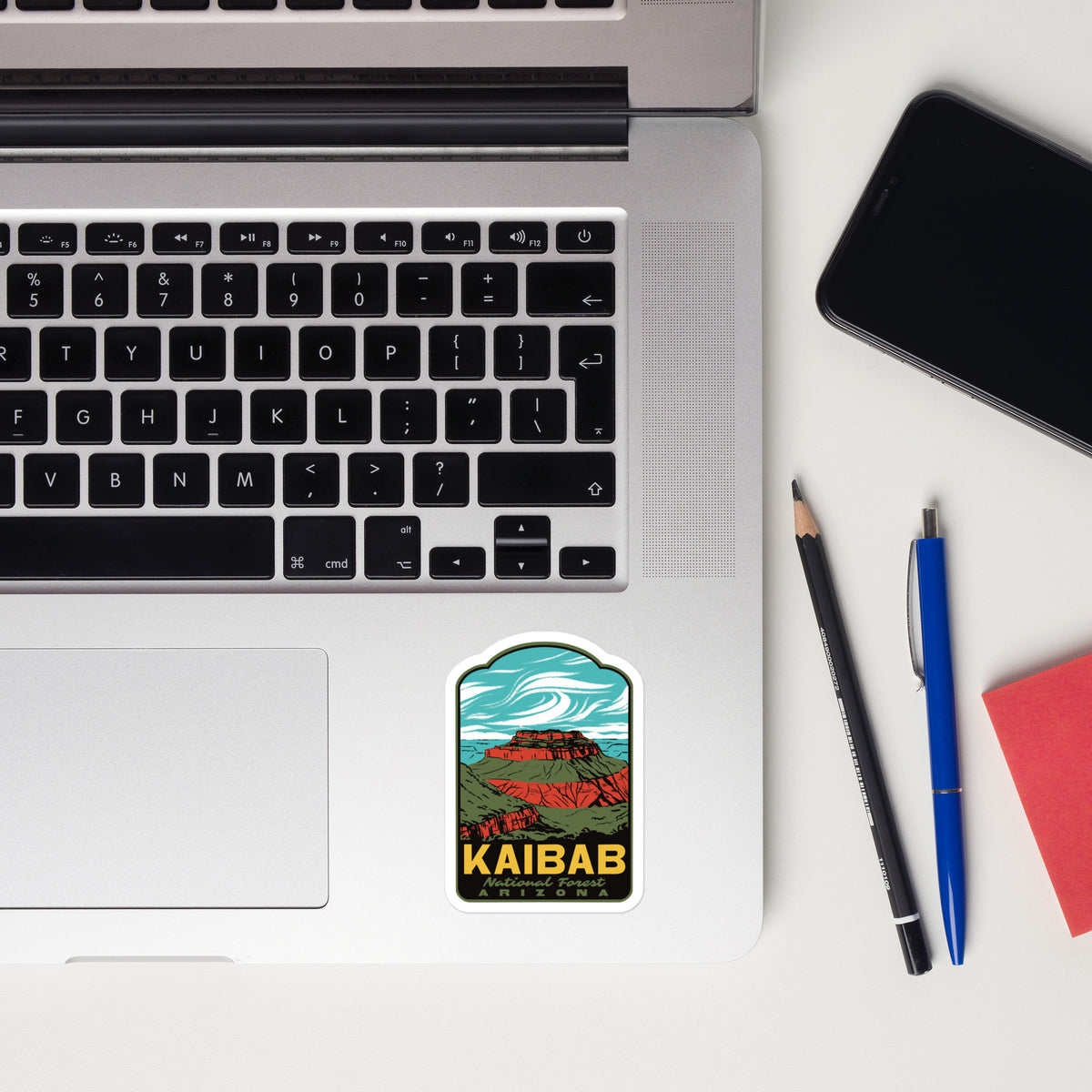 A rectangular sticker with rounded corners featuring a colorful landscape illustration and the text 'KAIBAB National Forest' is placed on the trackpad of an open silver laptop. The scene is styled as a flat lay on a white desk with a black smartphone, a pencil, a blue pen, and a red notebook.