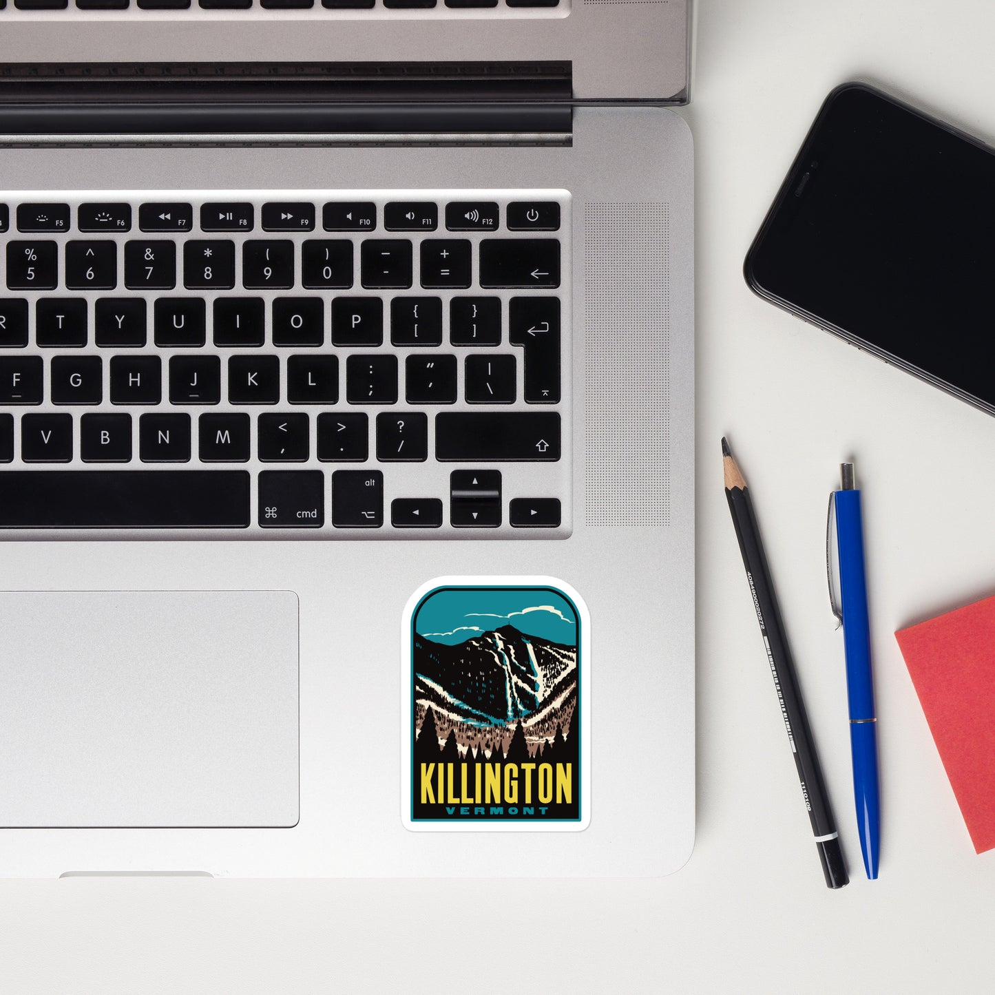 Laptop with a 'Killington Vermont' sticker, phone, pencil, and pen on a white surface