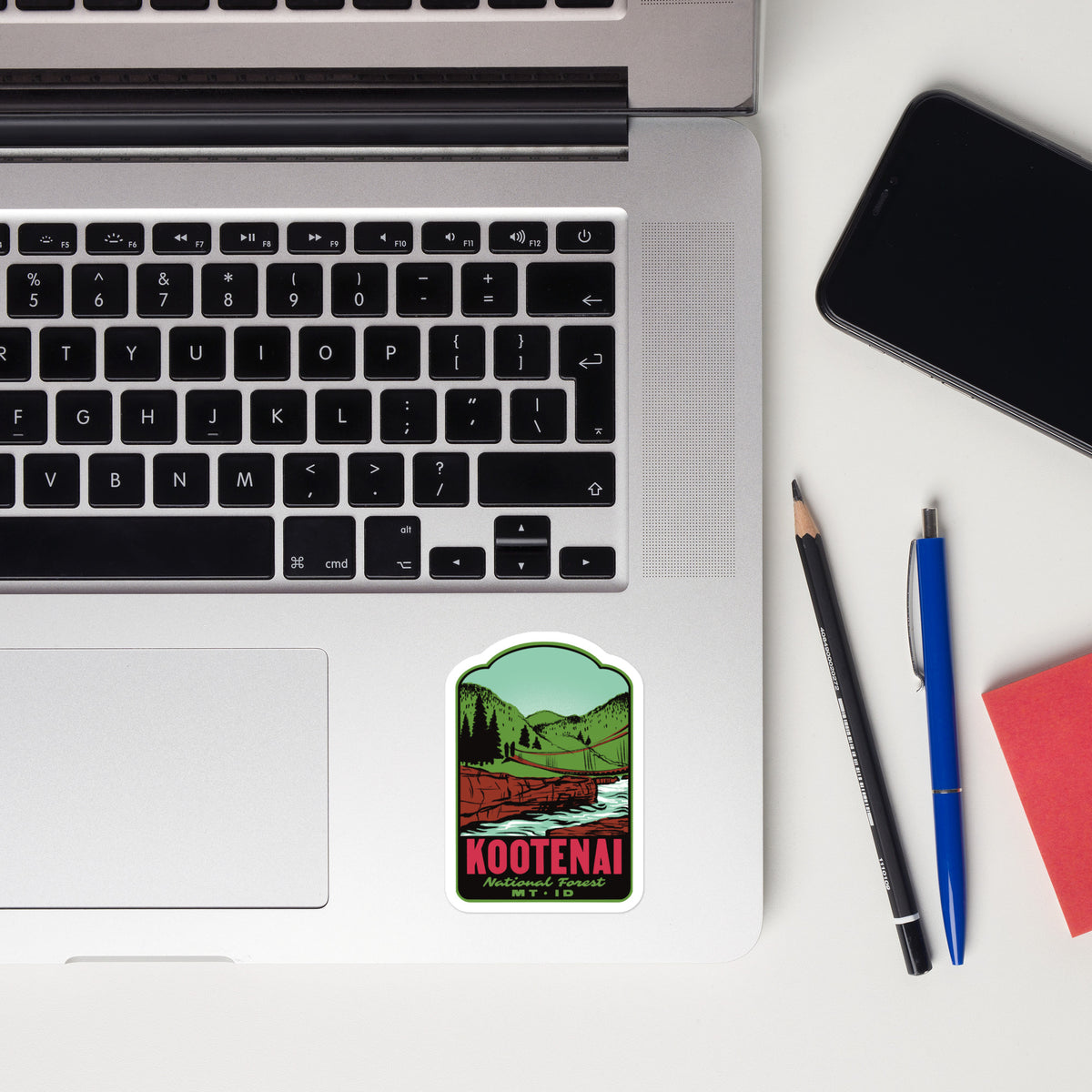 A rectangular sticker with rounded corners featuring a scenic green landscape and the text 'KOOTENAI' is placed on the palm rest of a silver laptop. The laptop is styled in a flat-lay arrangement alongside a smartphone, a pencil, a pen, and a red notebook on a white surface.