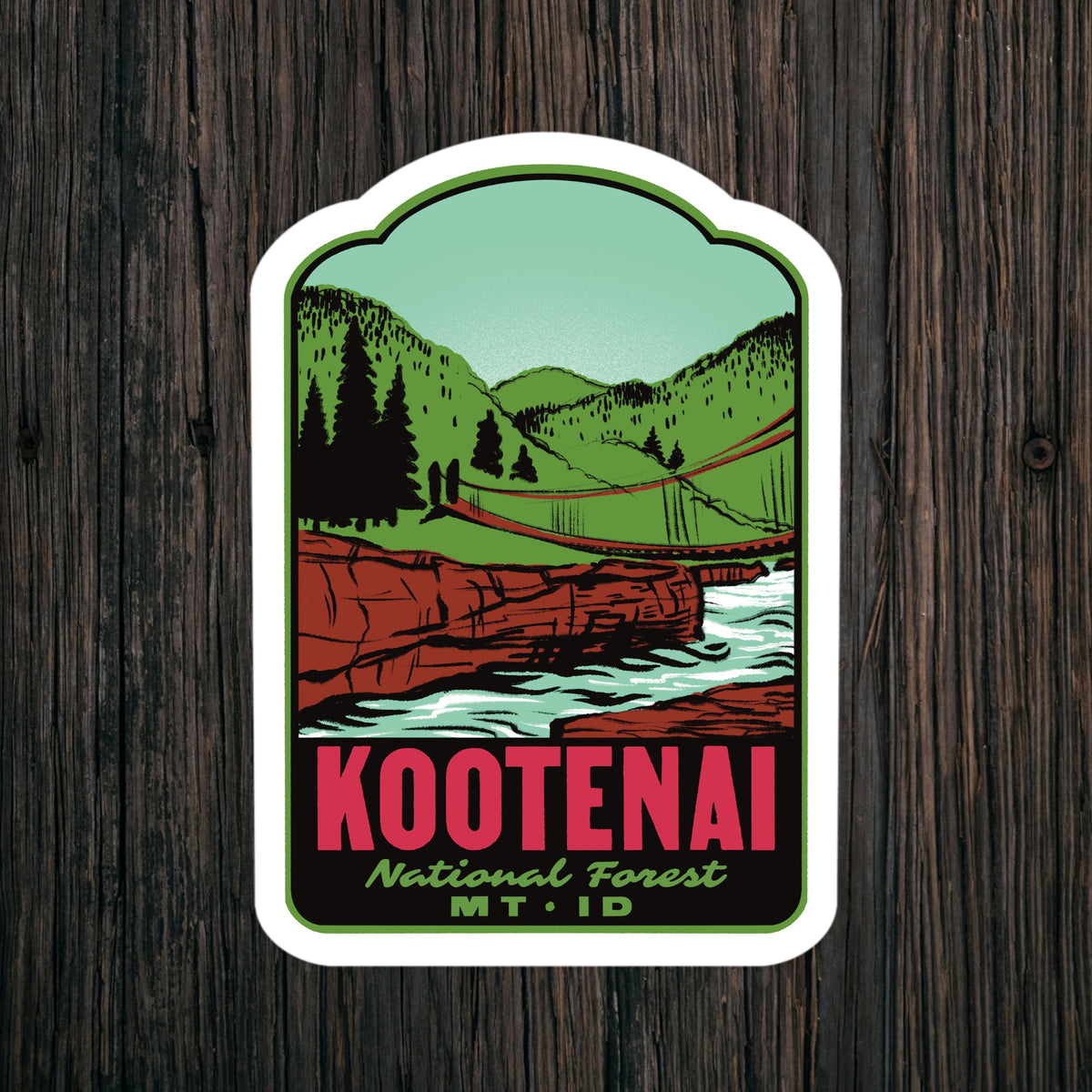A kiss-cut sticker featuring a vintage-style Kootenai National Forest illustration, displayed on a dark wood background. The sticker depicts green mountains, a red suspension bridge, a white river, and bold red text reading 'KOOTENAI' with 'National Forest MT.