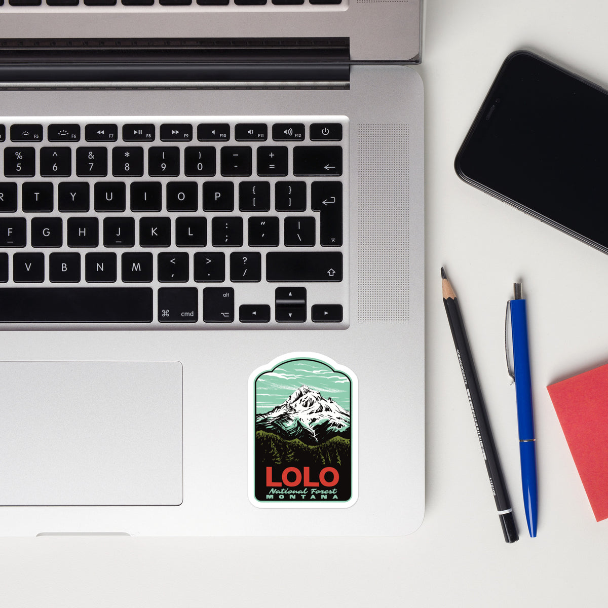 A rectangular sticker with rounded corners featuring a retro mountain illustration and the text 'LOLO' is shown on a silver laptop palm rest, styled in a flat-lay with a smartphone, pencil, pen, and red notebook on a white desk.