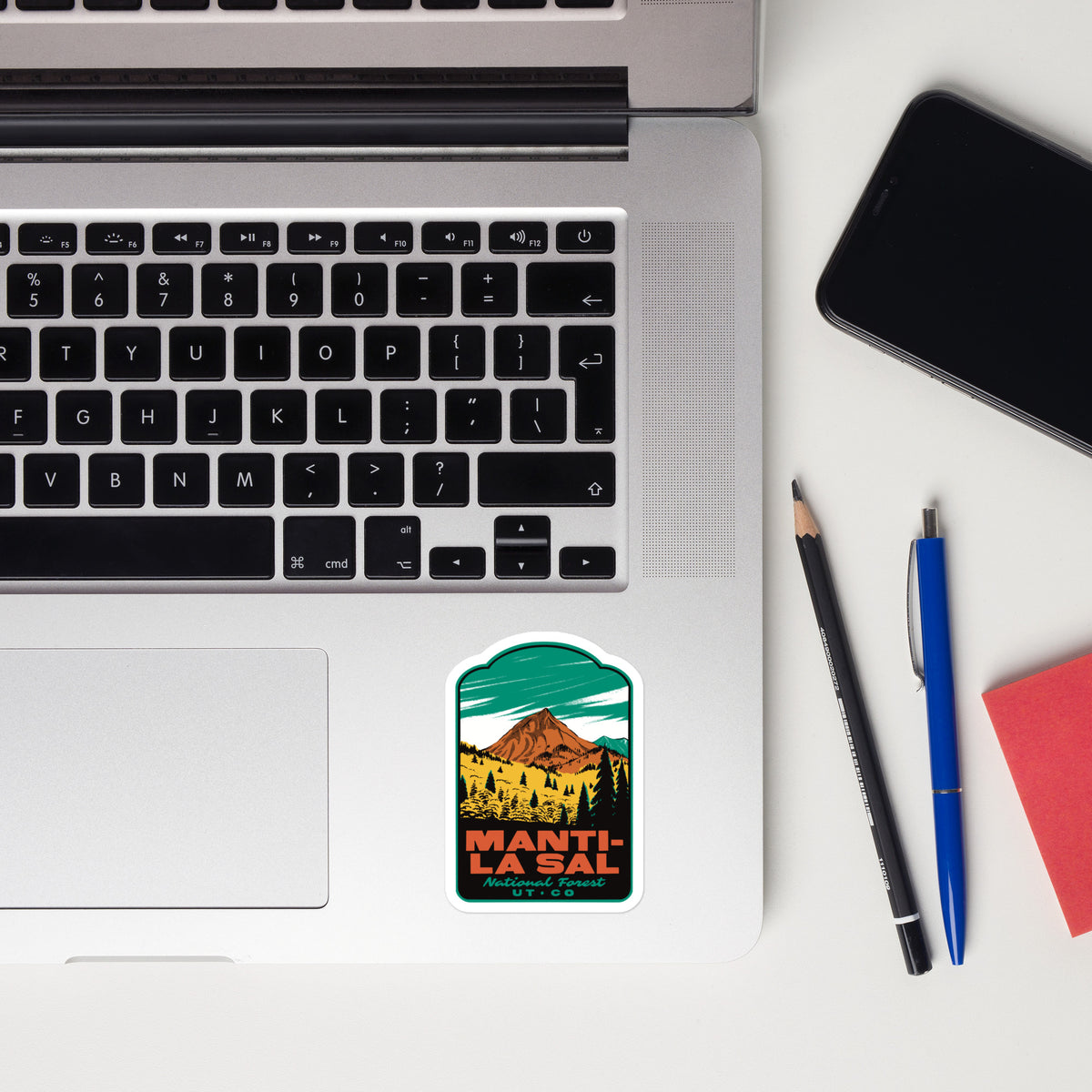 A rectangular sticker with rounded corners featuring a retro mountain illustration and the text 'MANTI-LA-SAL' is placed on the lower right corner of a silver laptop keyboard. The laptop sits on a white desk alongside a black smartphone, a pencil, a blue pen, and a red notebook.