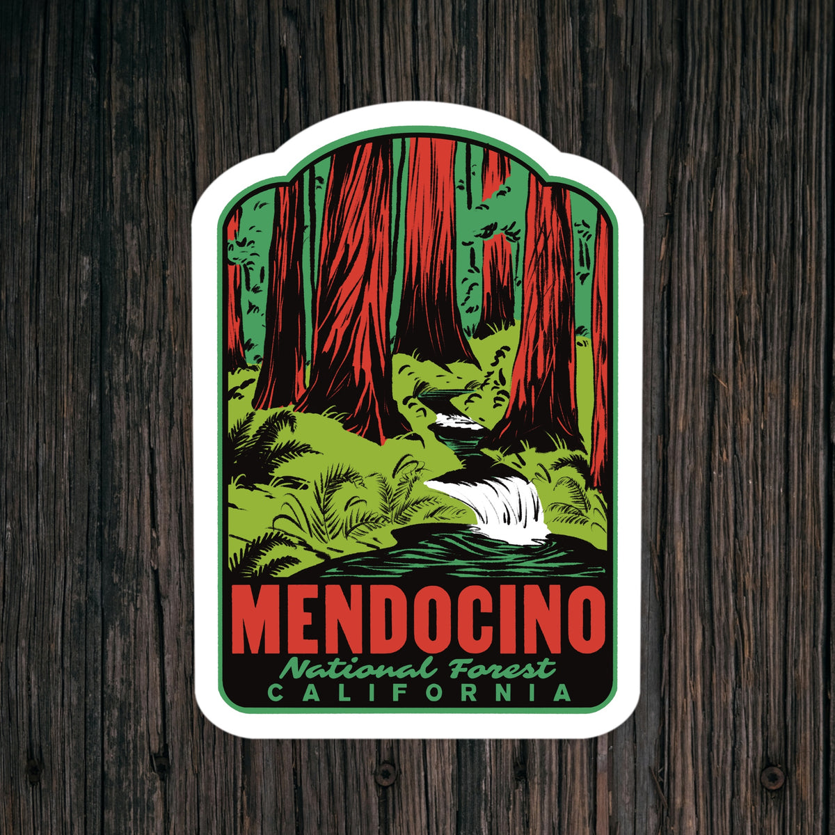 A die-cut sticker featuring a vintage-style illustration of a redwood forest and waterfall with the text 'MENDOCINO National Forest CALIFORNIA', shown against a dark wood plank background.