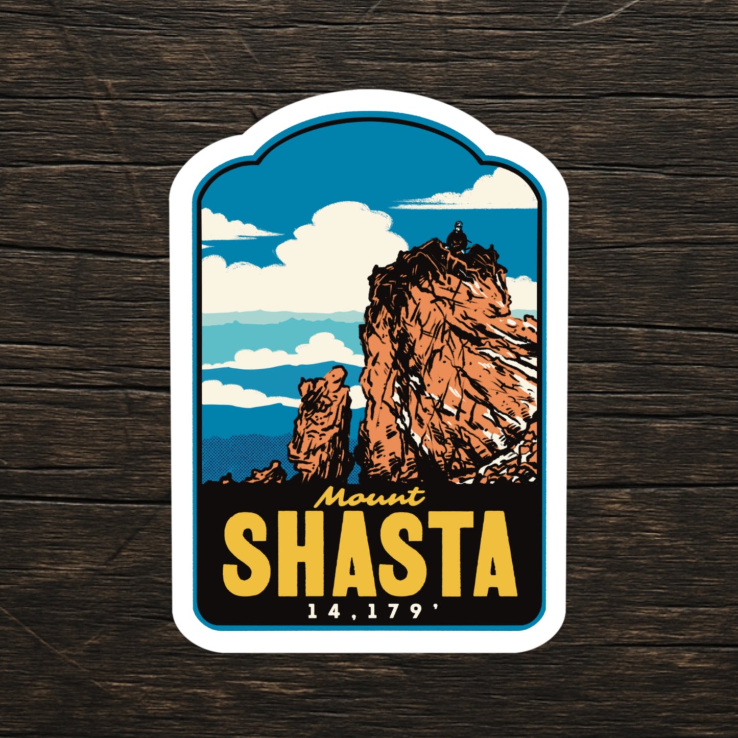 A die-cut sticker featuring a retro-style illustration of Mount Shasta, displayed on a dark wood background. The sticker has a white border and displays the text 'Mount SHASTA 14,179' in yellow and white lettering.