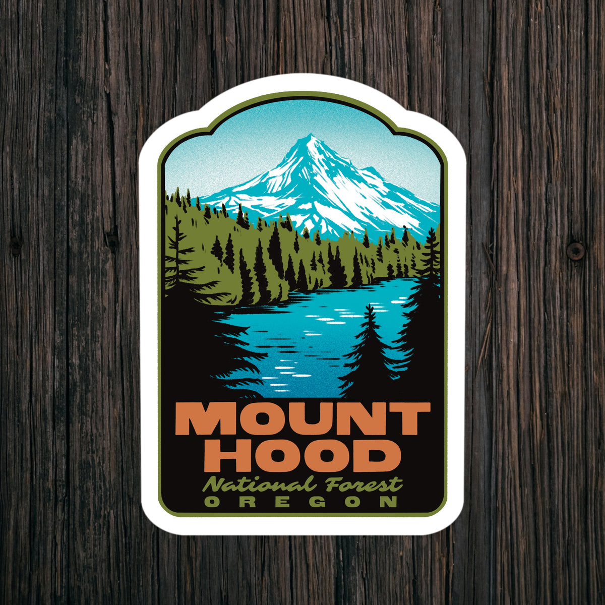 A die-cut sticker featuring a retro-style illustration of Mount Hood National Forest in Oregon, with a snowy mountain, blue lake, pine trees, and bold orange text, shown against a dark wood plank background.