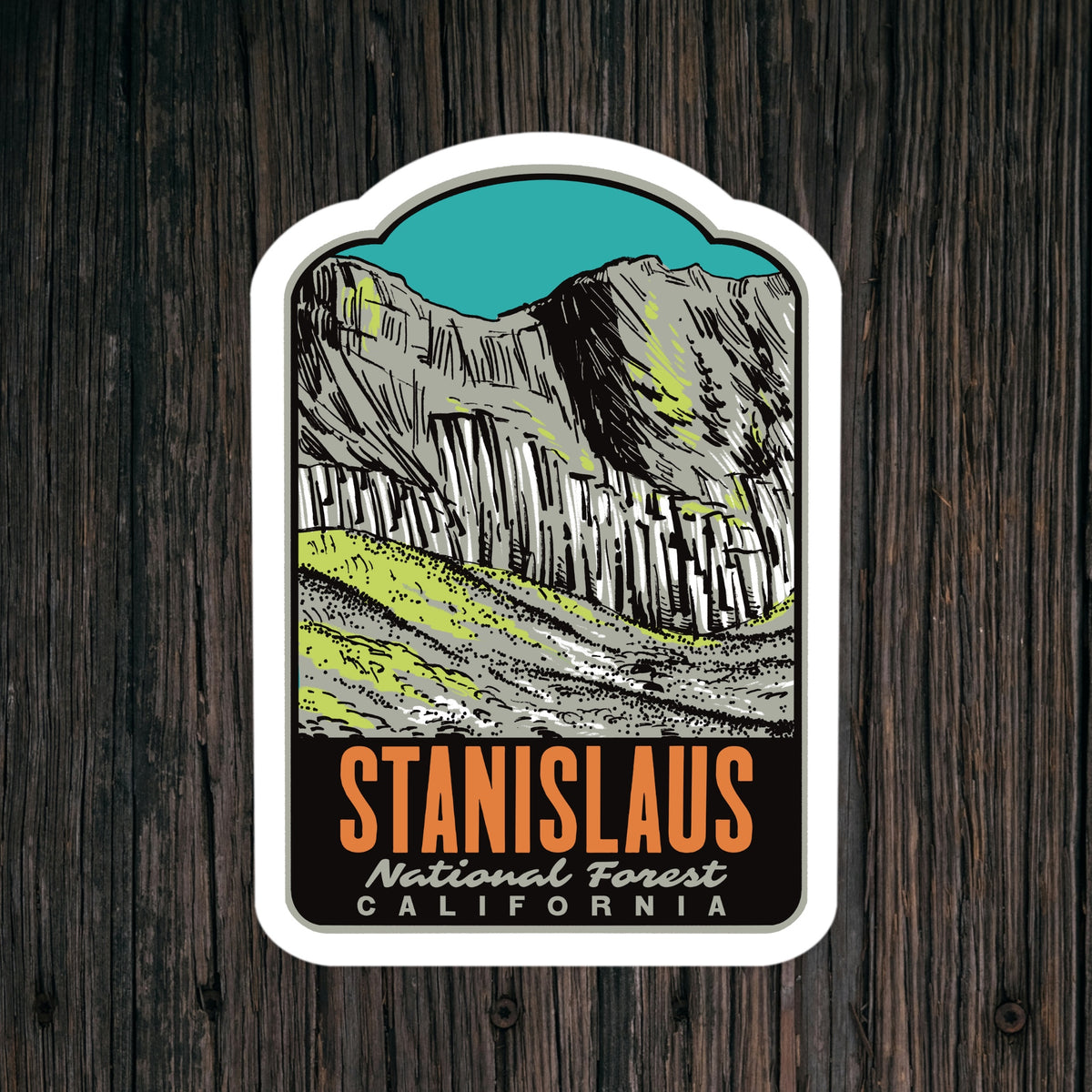 A kiss-cut sticker featuring an illustration of Stanislaus National Forest in California, displayed on a dark wood background. The sticker has a white border and displays the text 'STANISLAUS National Forest CALIFORNIA' in orange and white lettering.