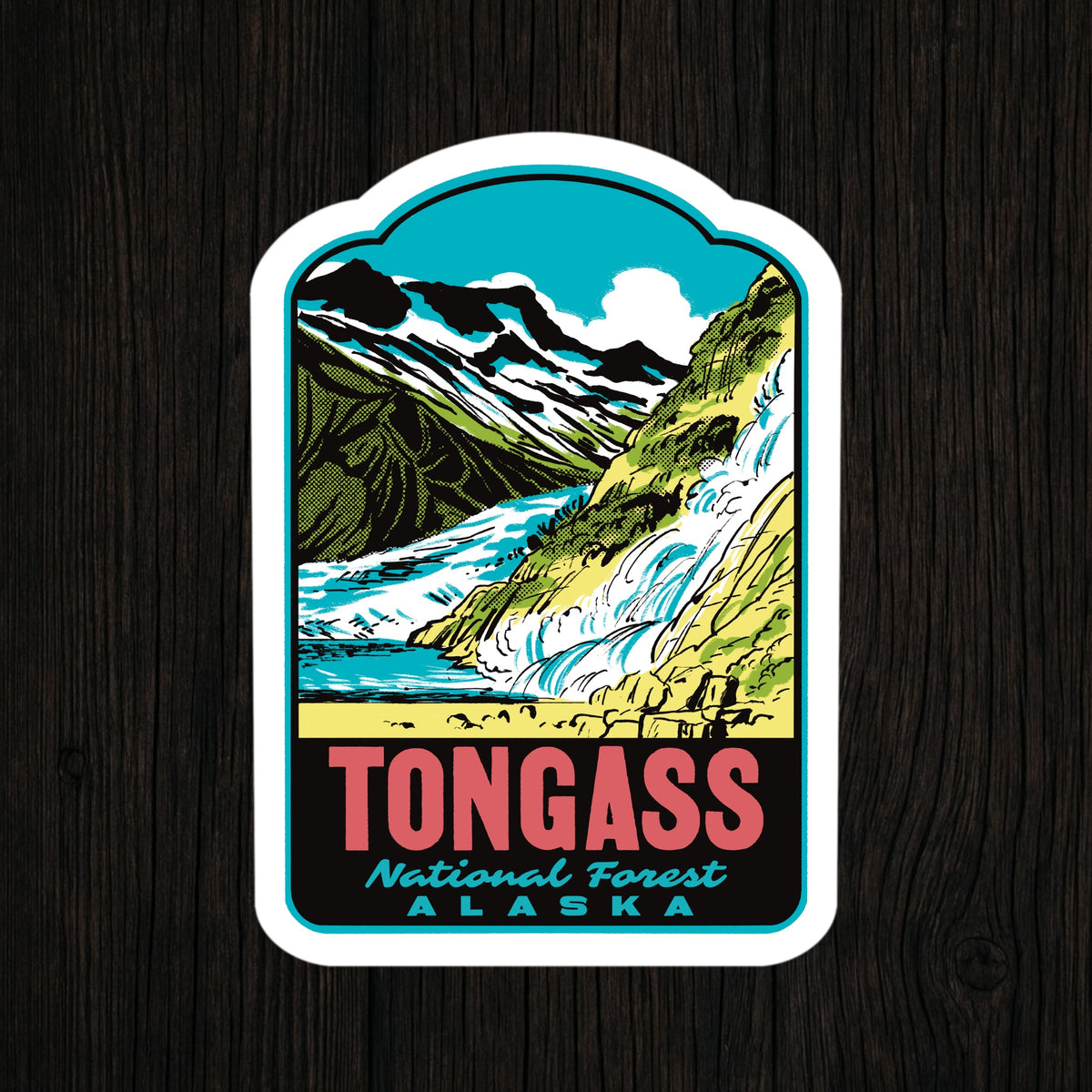 A die-cut sticker featuring a stylized illustration of Tongass National Forest in Alaska, with mountains, a waterfall, and the text 'TONGASS National Forest ALASKA,' shown on a dark wood background.