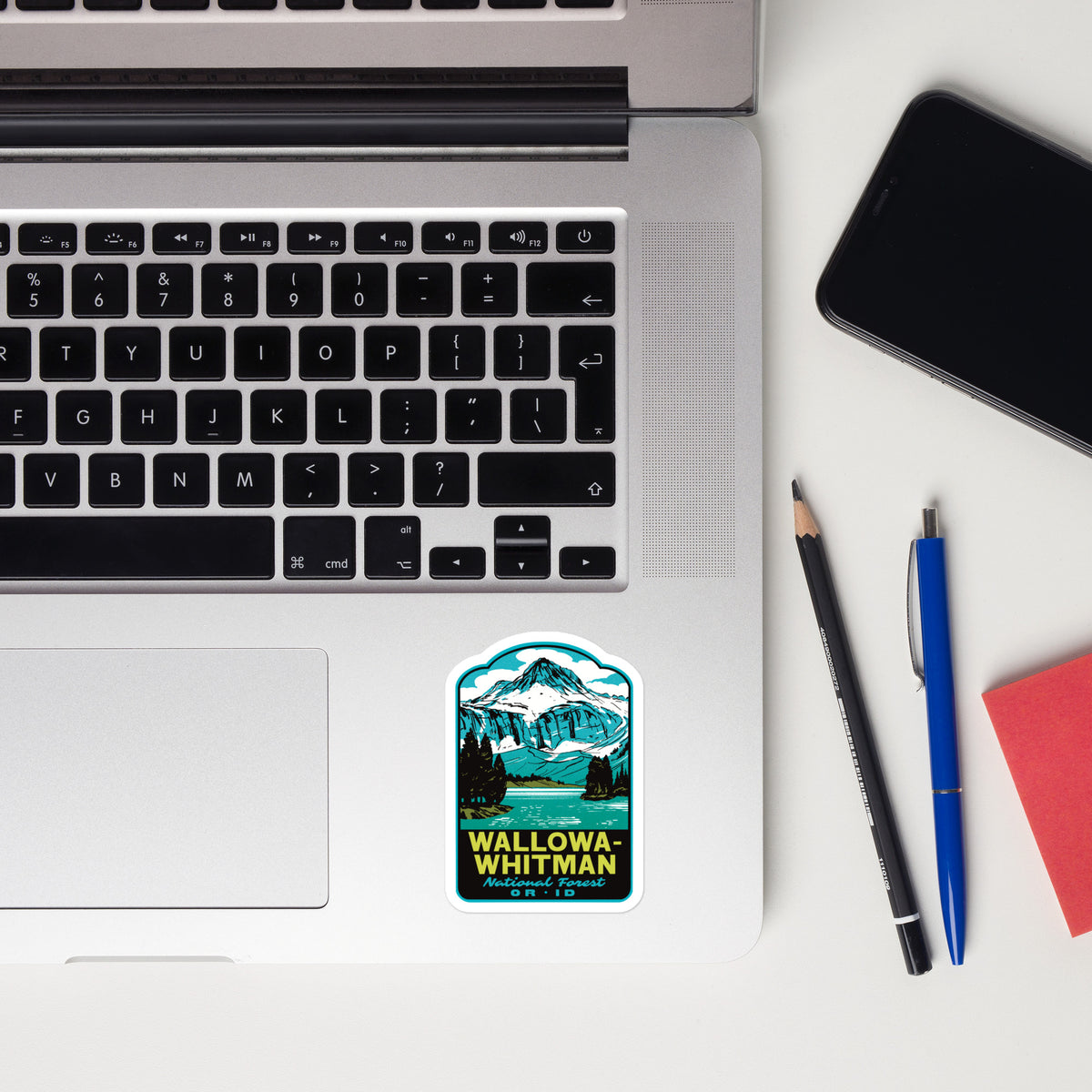 A rectangular sticker with a mountain landscape illustration and the text 'WALLOWA-WHITMAN' is placed on the palm rest of an open silver laptop. The scene is styled as a flat lay on a white desk with a black smartphone, a pencil, a blue pen, and a red notebook nearby.