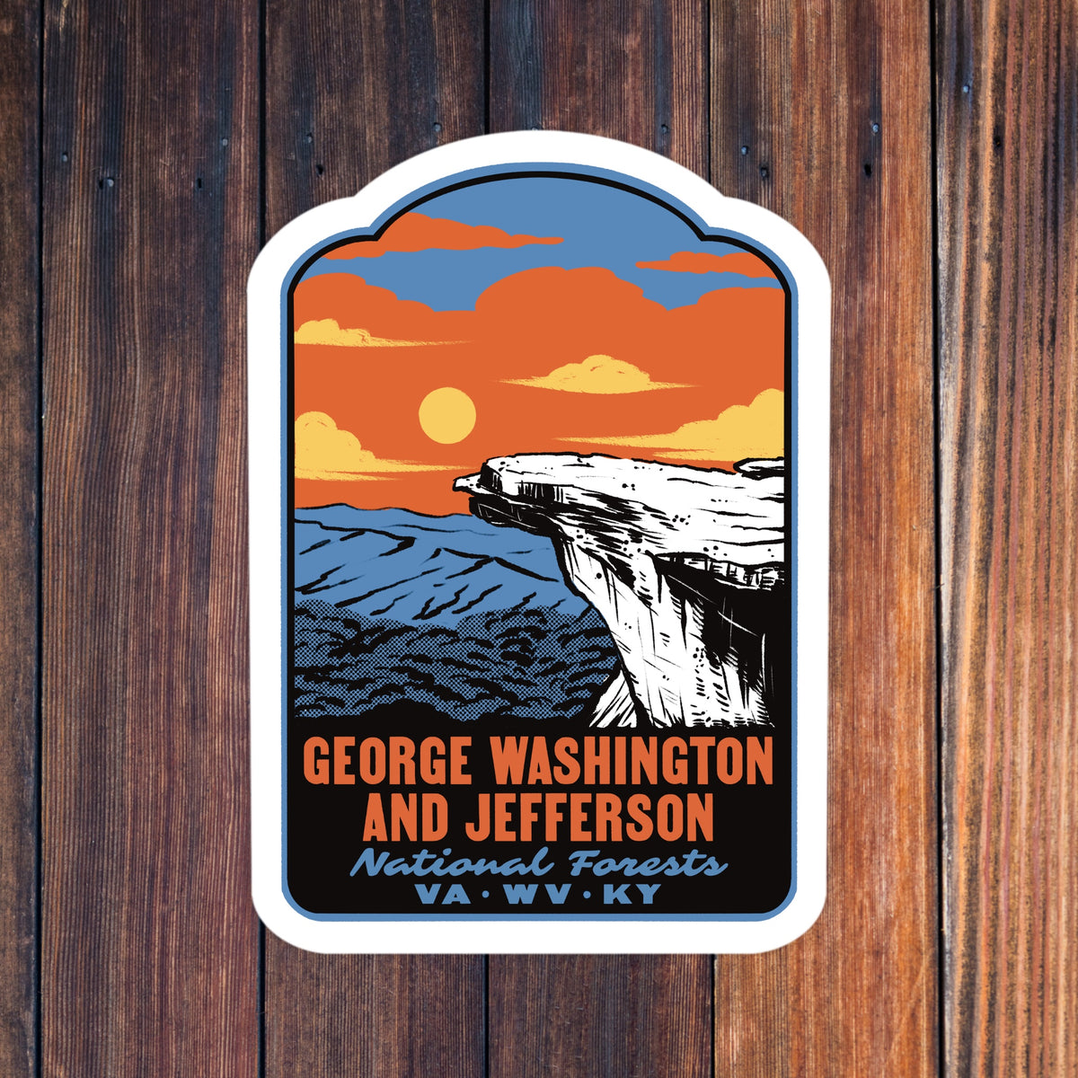 A die-cut sticker featuring a retro-style landscape illustration of a cliff and sunset, with the text 'GEORGE WASHINGTON AND JEFFERSON National Forests VA·WV·KY', shown against a dark wood plank background.