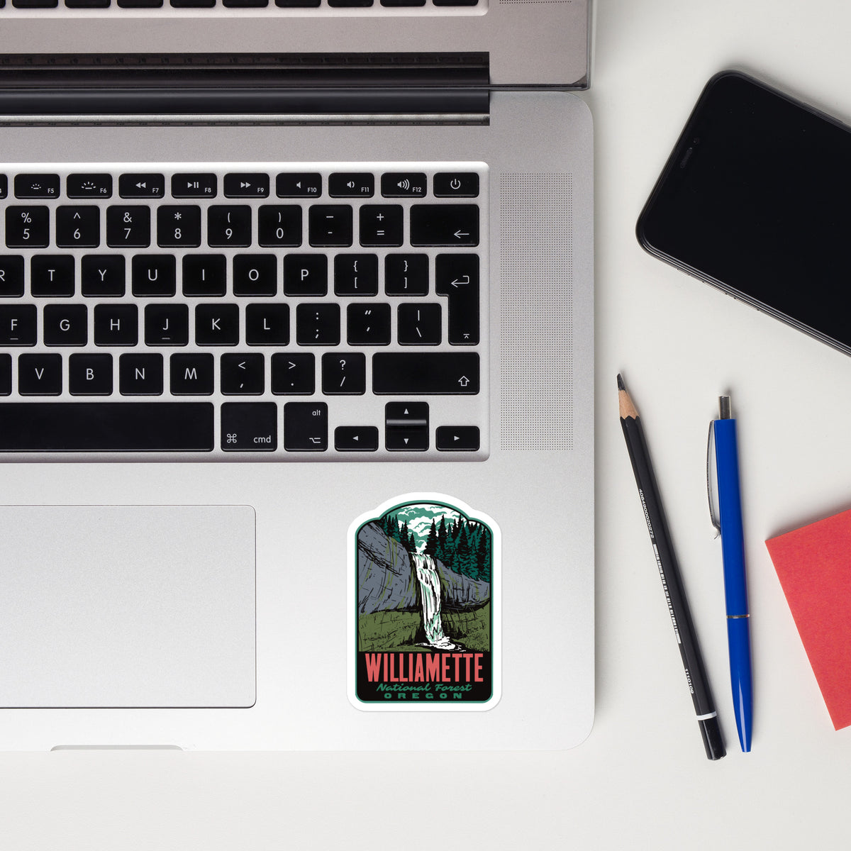 A rectangular die-cut sticker featuring a stylized illustration of a woman in a white dress standing in front of a dark mountain landscape, with the word 'WILLIAMETTE' in red at the bottom. The sticker is placed on the lower-left corner of an open silver laptop on a white desk, alongside a black smartphone, a pencil, a blue pen, and a red notebook.