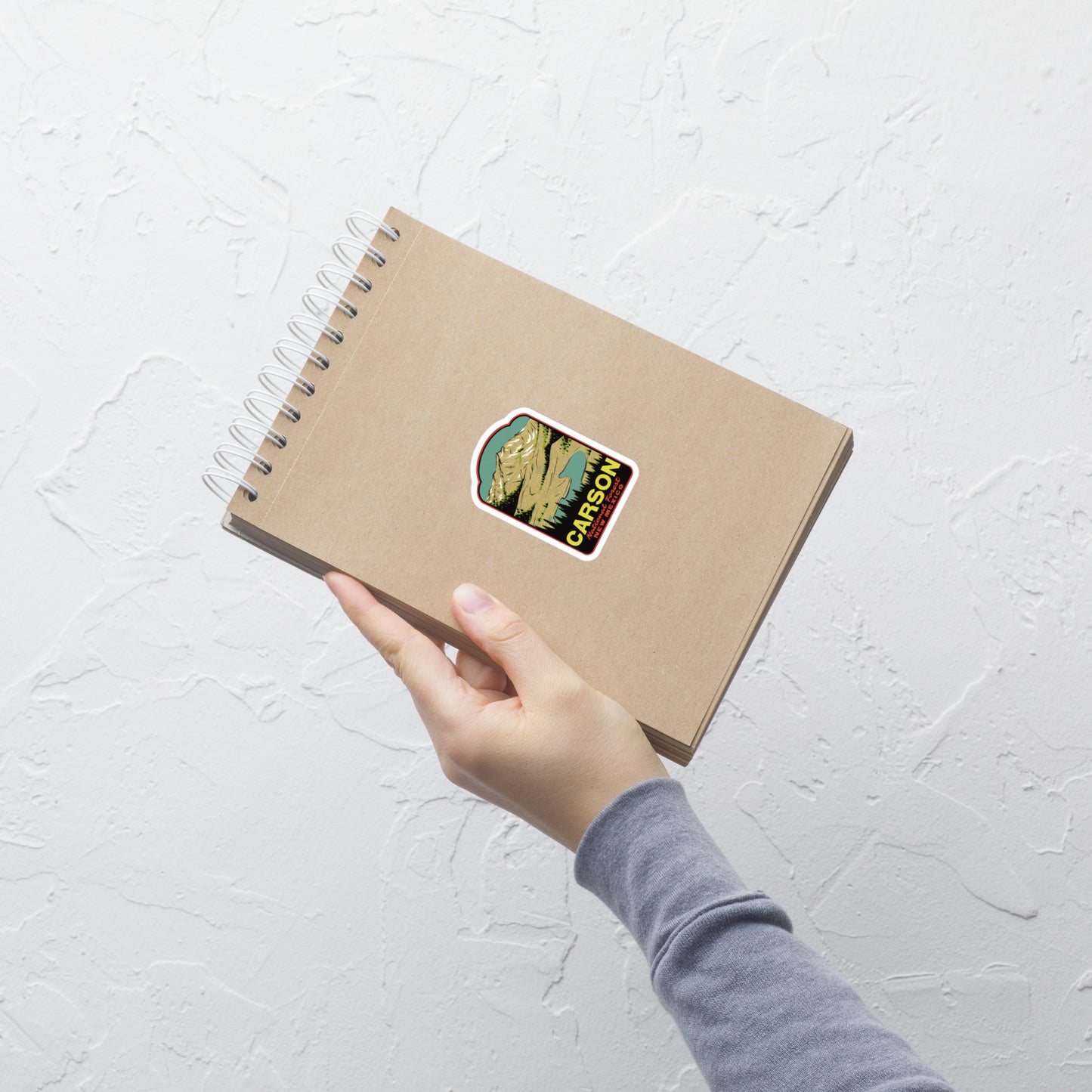 Hand holding a brown notebook with a Carson National Forest sticker on a light gray textured background