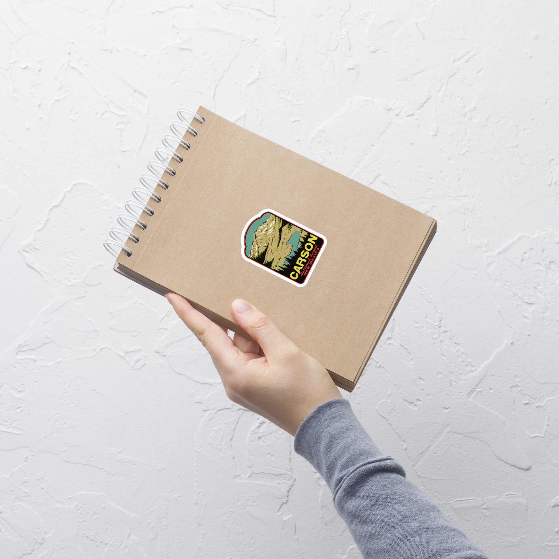 Hand holding a brown notebook with a Carson National Forest sticker on a light gray textured background