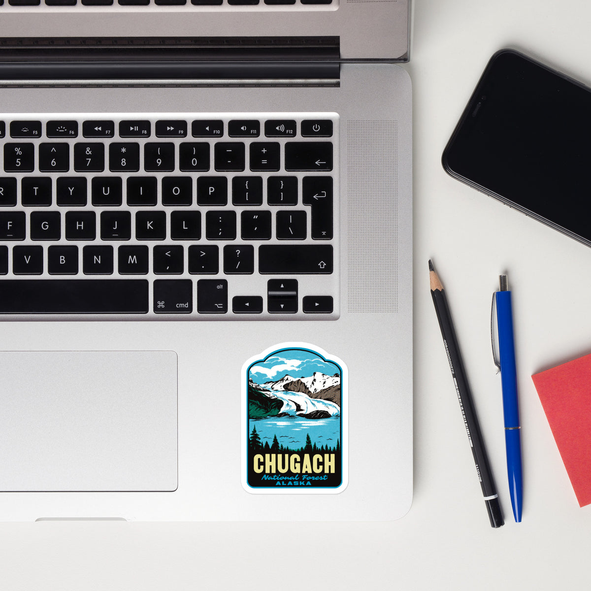 A rectangular sticker with rounded corners featuring a mountain and lake illustration and the text 'CHUGACH ALASKA' is placed on the lower right corner of a silver laptop. The laptop is shown in a styled flat-lay on a white desk alongside a smartphone, a pencil, a pen, and a red notebook.