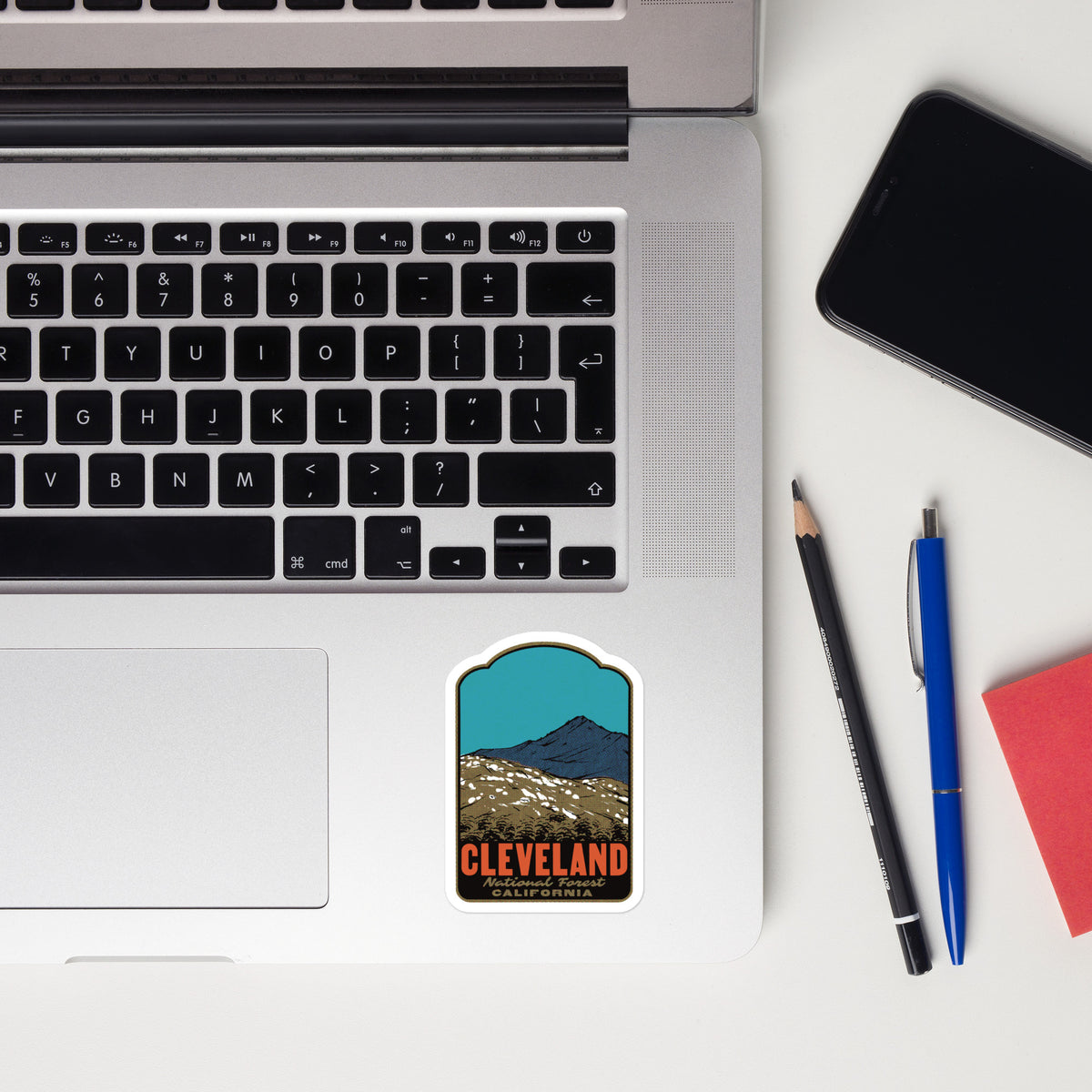 A rectangular sticker with an arched top featuring a retro-style illustration of Cleveland, Ohio, placed on the bottom right corner of a silver laptop. The sticker depicts a blue sky, a dark mountain peak, and a brown landscape with the word 'CLEVELAND' in orange.