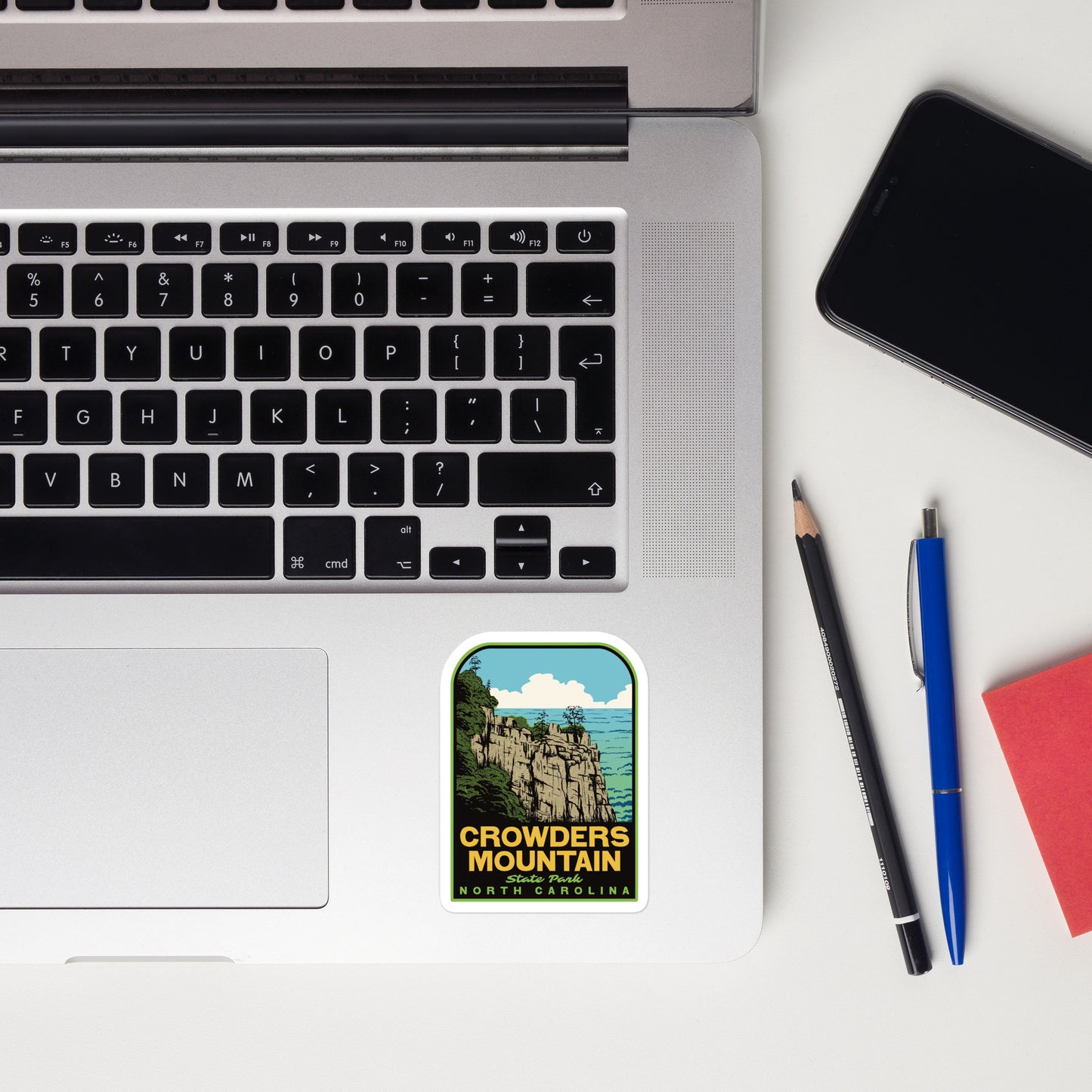 Laptop with a 'Crowders Mountain, North Carolina' sticker, phone, pencil, pen, and notebook on a white surface.