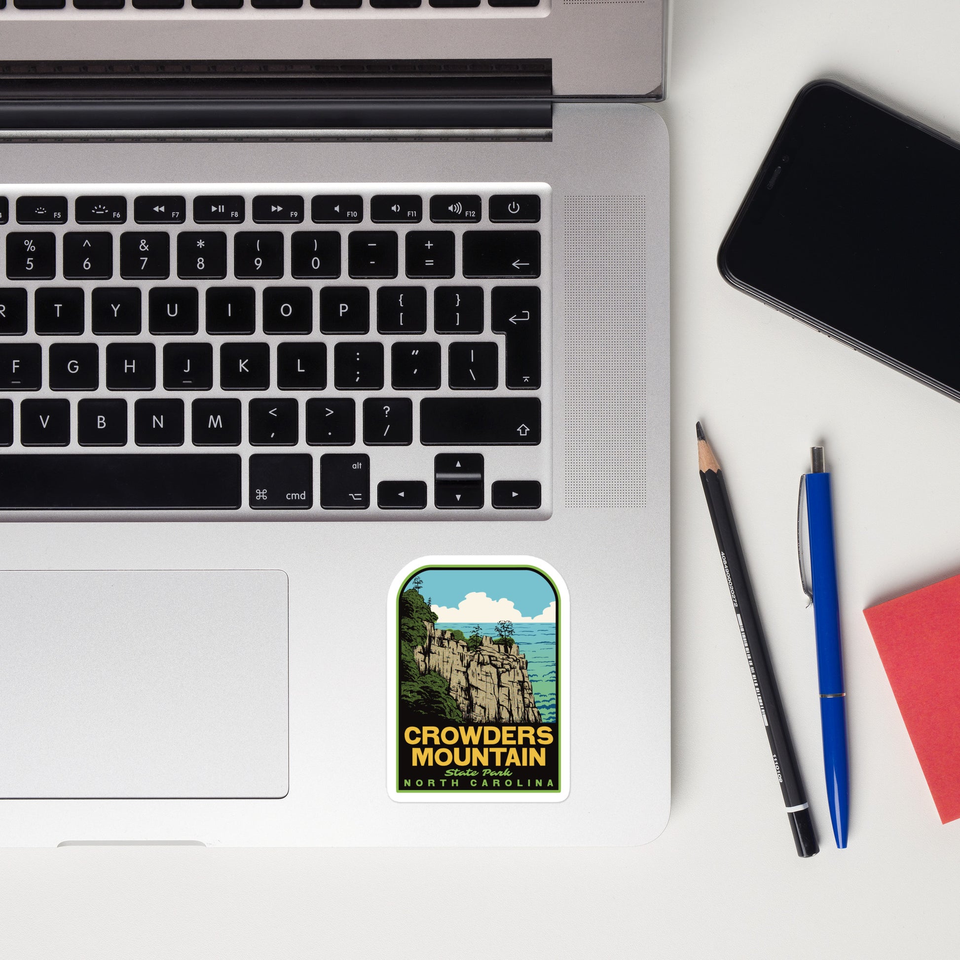 Laptop with a 'Crowders Mountain, North Carolina' sticker, phone, pencil, pen, and notebook on a white surface.
